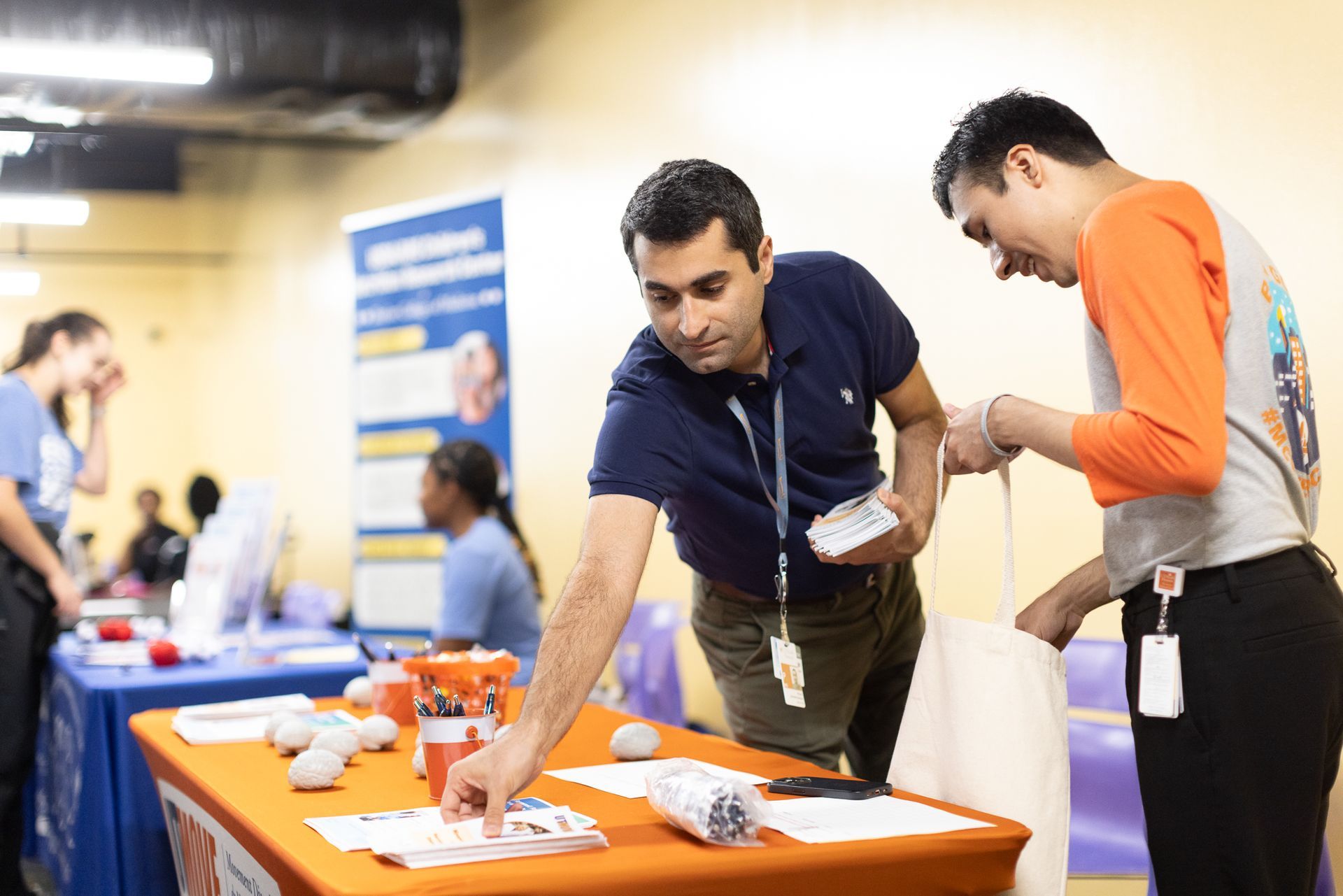 Two men at a table, one handing a bag. Table has flyers and items. Others in background.