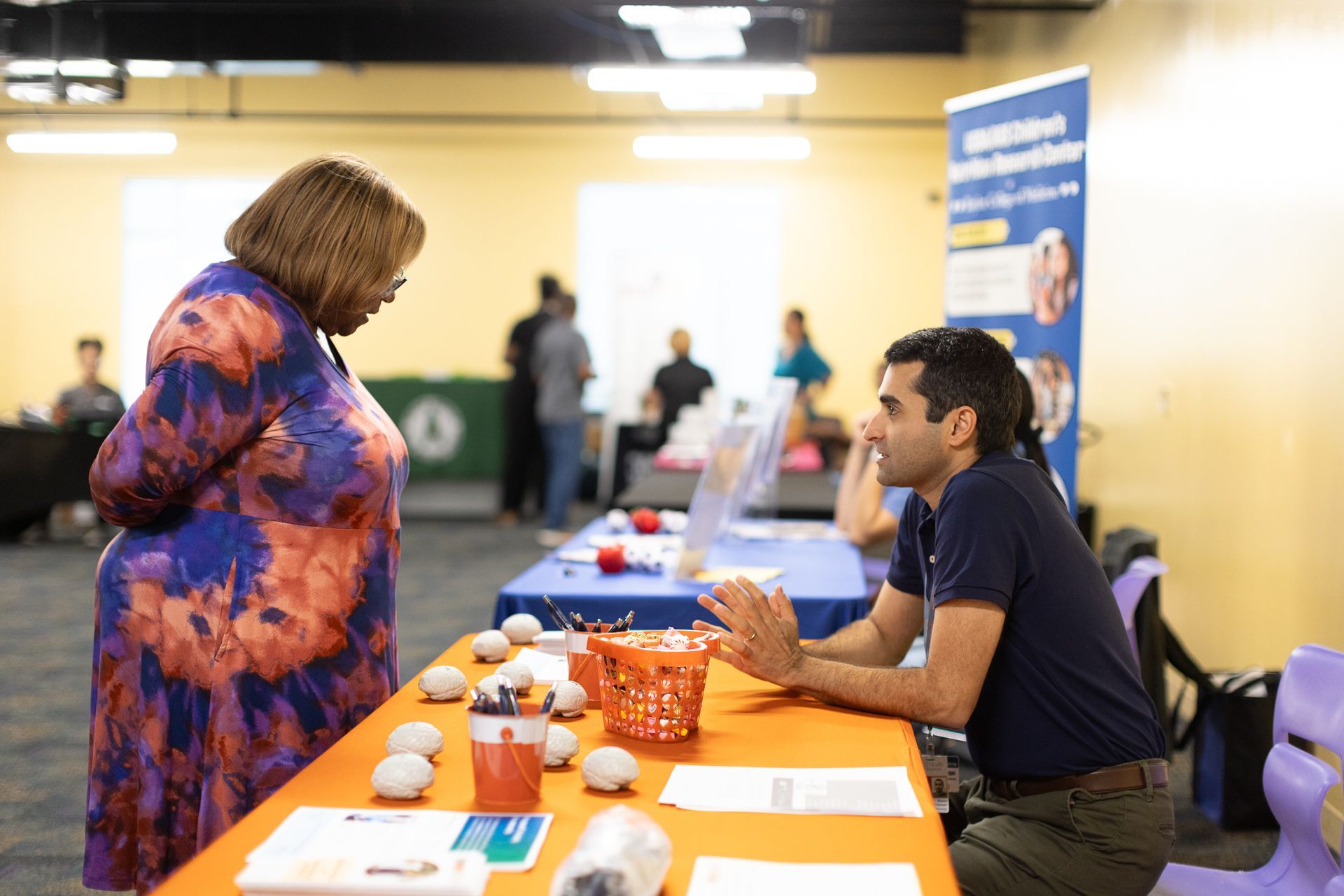 Woman speaks with a man at a table, likely an event. Others in background.