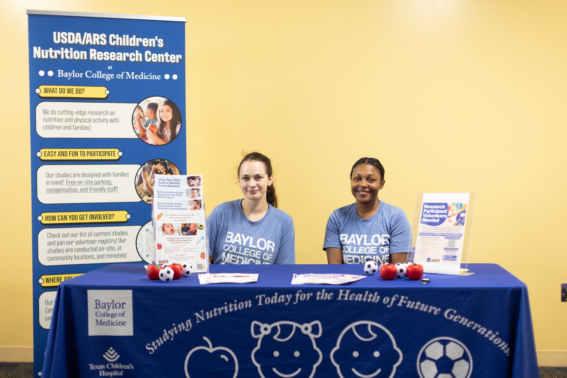 Two women at a Baylor Children's Nutrition Research Center booth with blue tablecloth and banner.