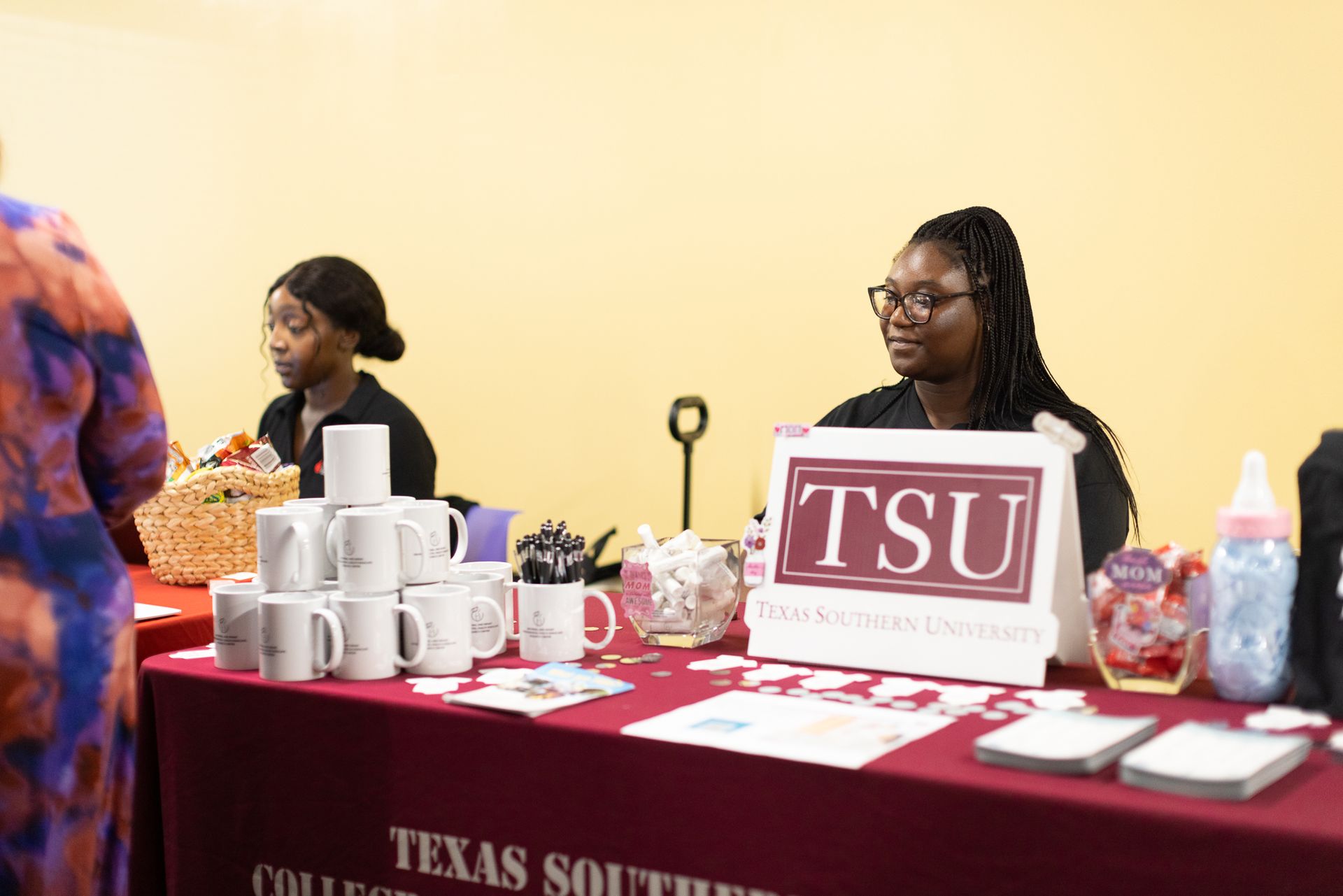 Two people at a Texas Southern University information booth; maroon table with mugs, sign, brochures.