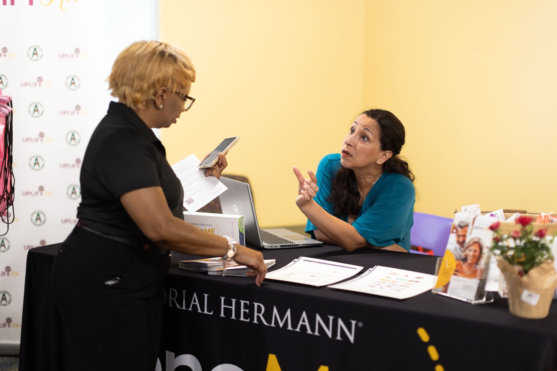 Woman at a table with another woman, both looking at paperwork; a logo on the table.