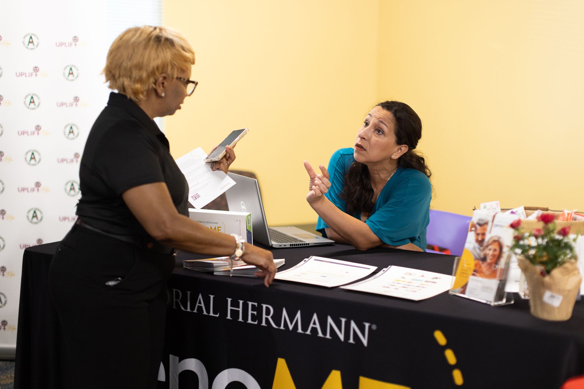 Two women at a Memorial Hermann information table, one is holding a phone and paperwork, the other is gesturing, speaking.