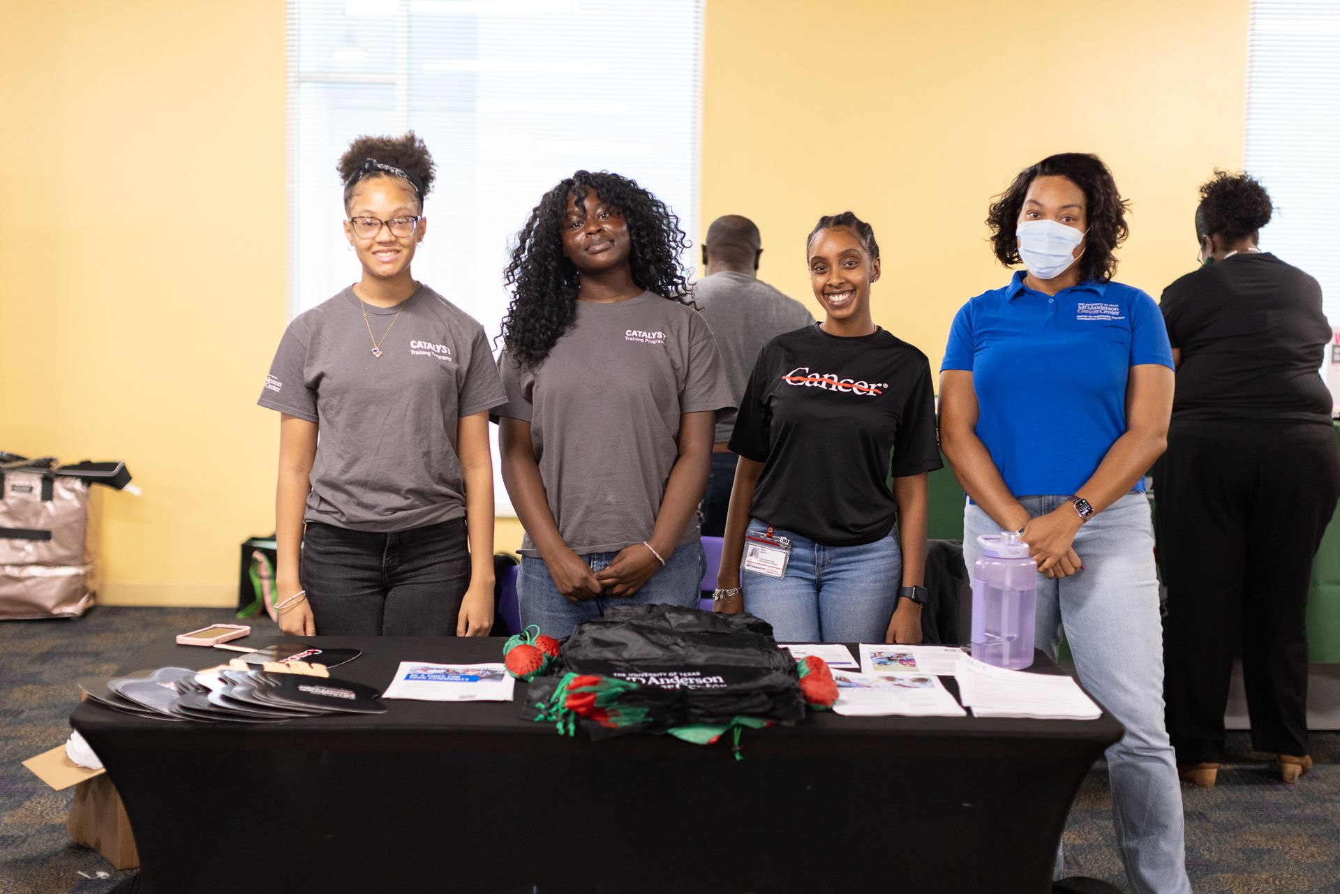 Four people at a table; they are smiling and wearing t-shirts and a mask, in front of a yellow wall.