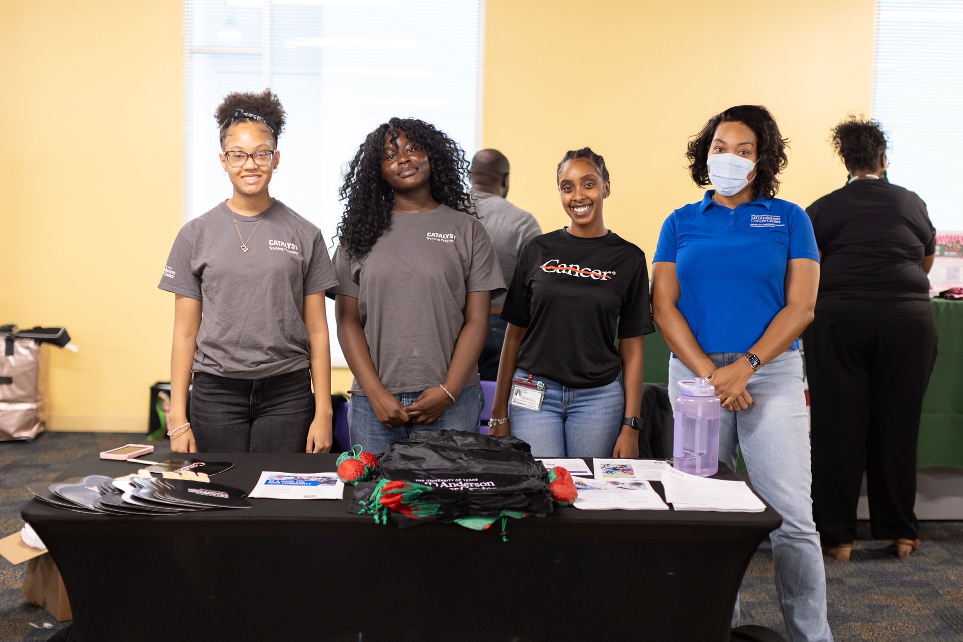Four people stand behind a table, smiling. Items and papers on the table. Yellow wall in the background.