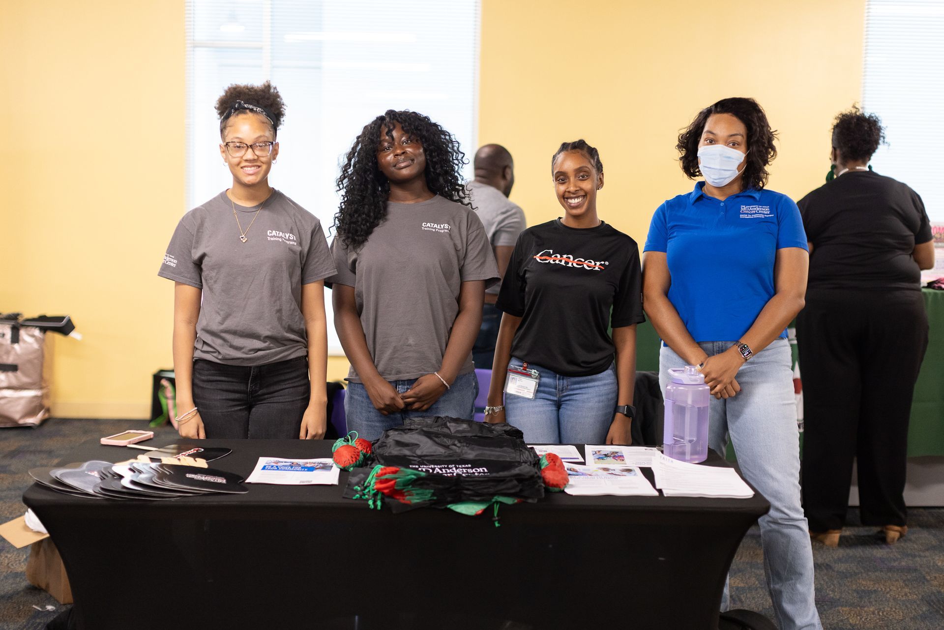 Four people at a table; one wearing a mask. They are smiling, with promotional items visible. Indoors.