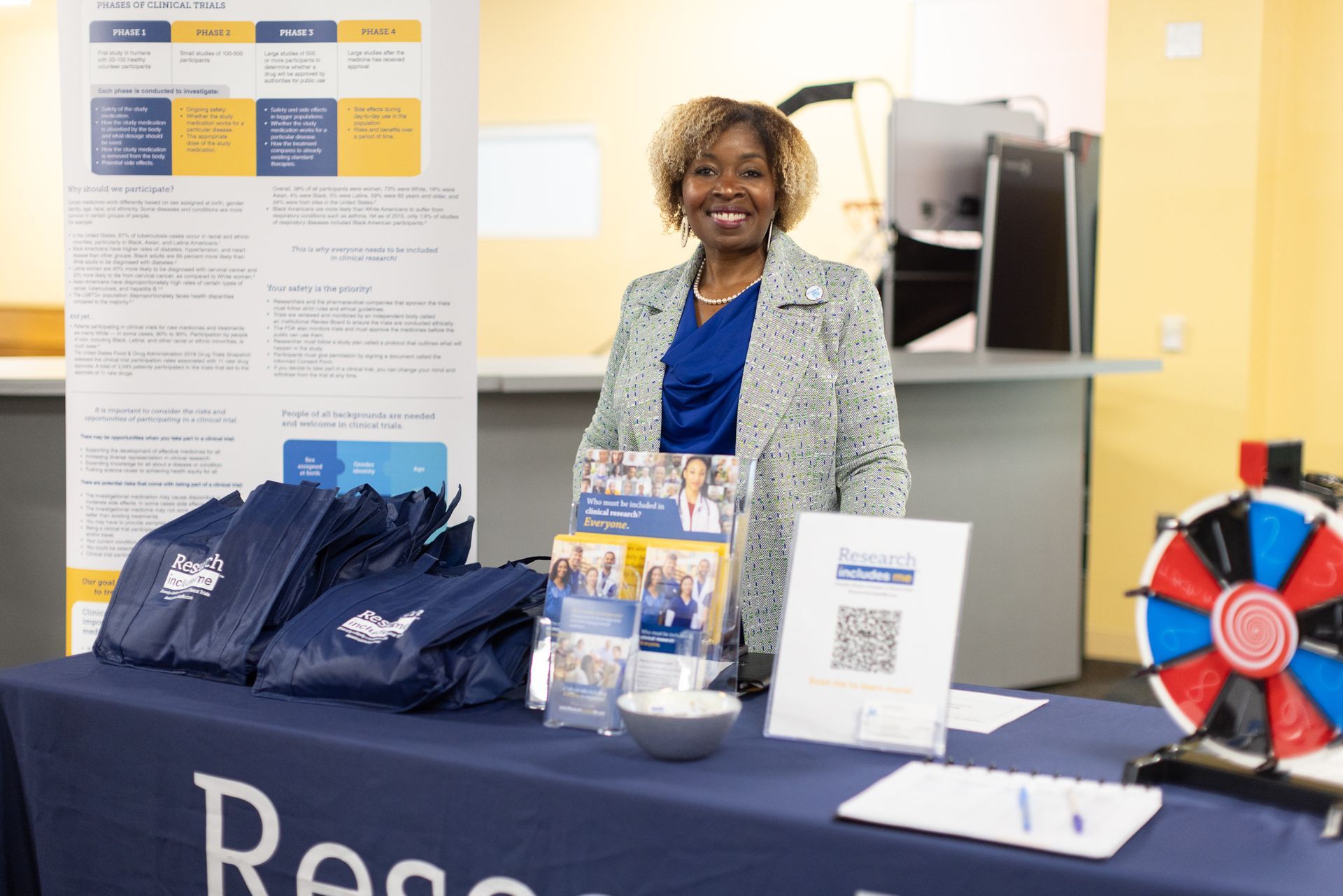 Woman at a table with promotional materials, smiling, standing in front of a banner.