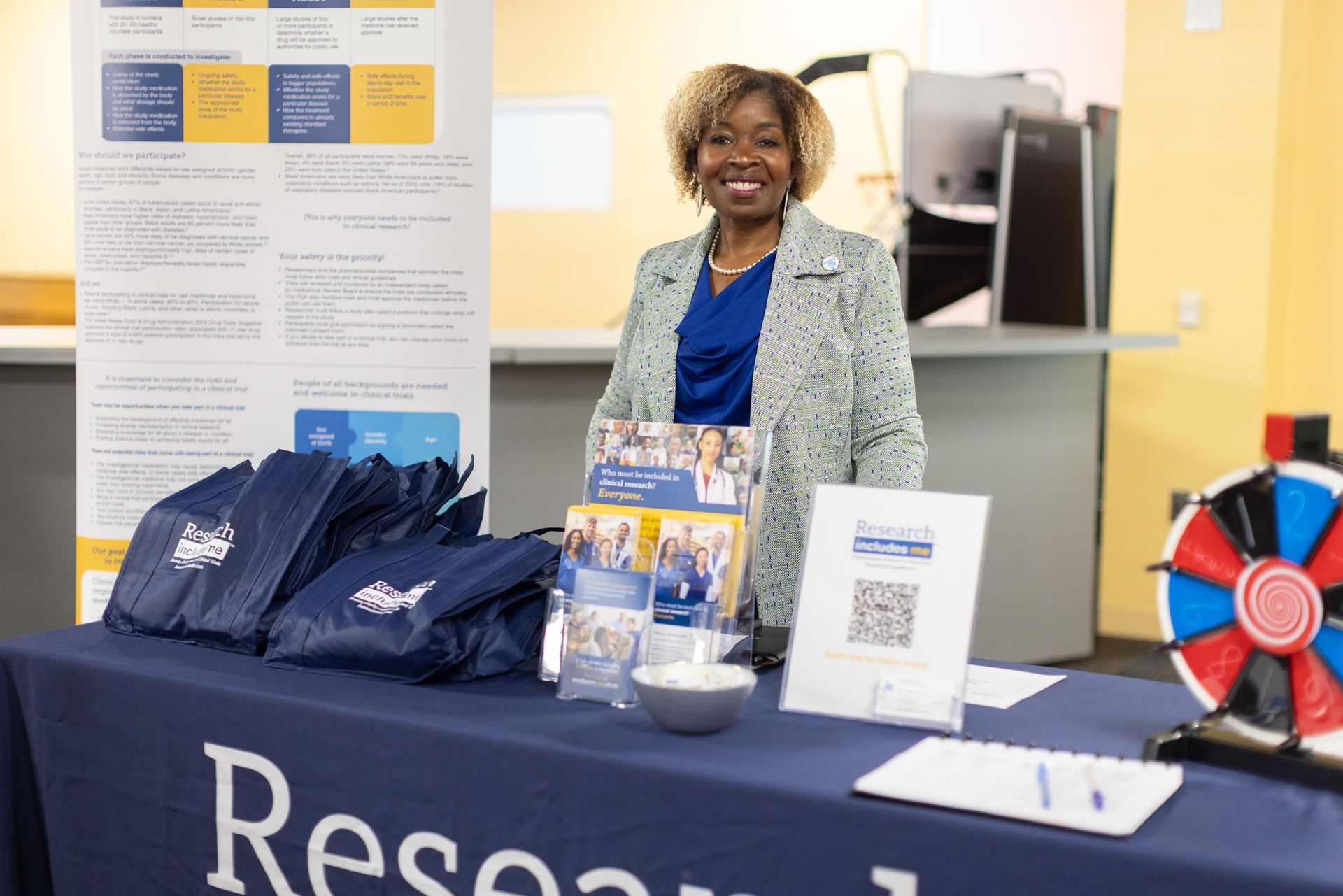 Woman at a table with promotional materials. Blue and yellow colors. She's smiling, standing behind bags and a prize wheel.