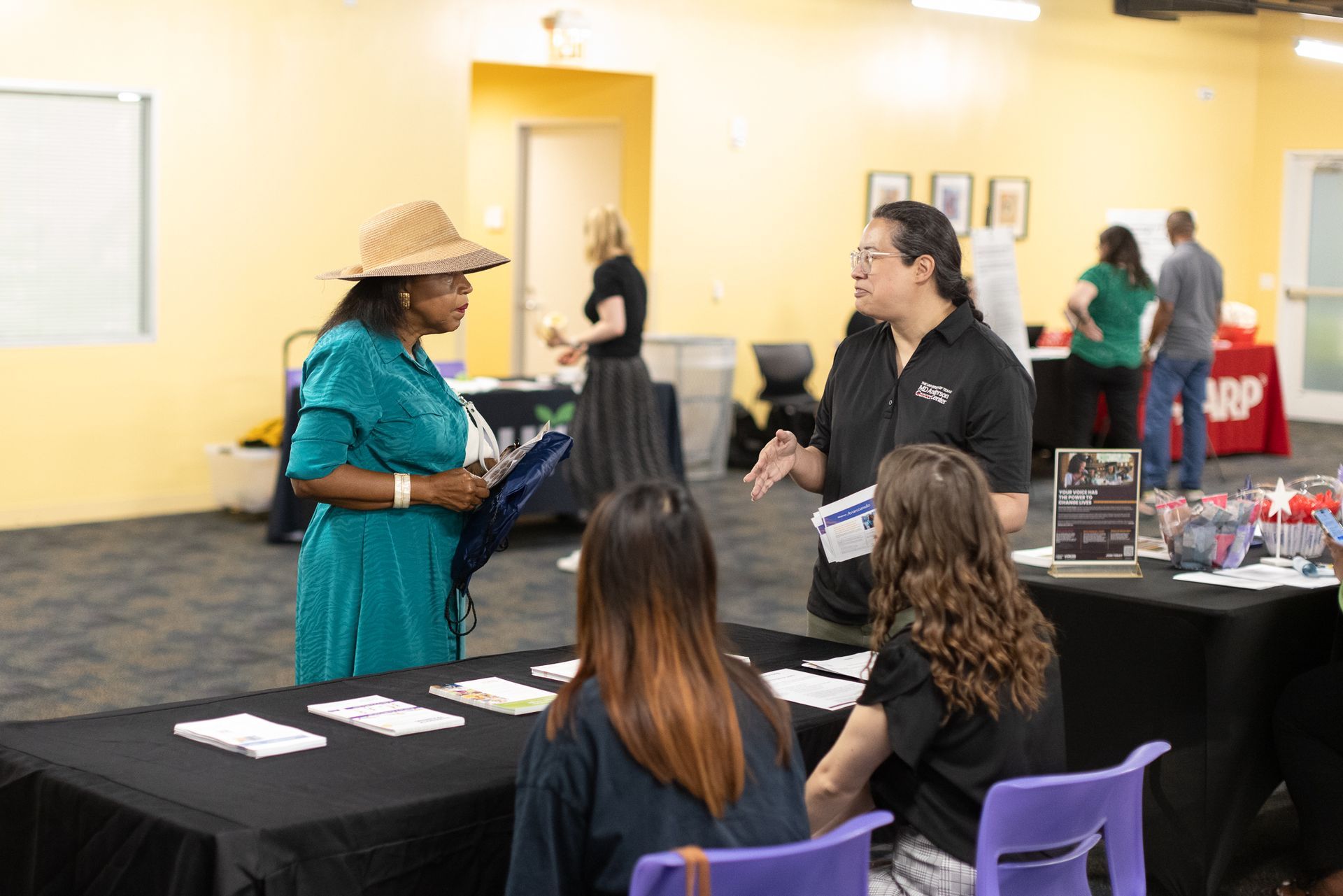People at a booth, discussing, at an event with tables and other attendees in a brightly lit room.