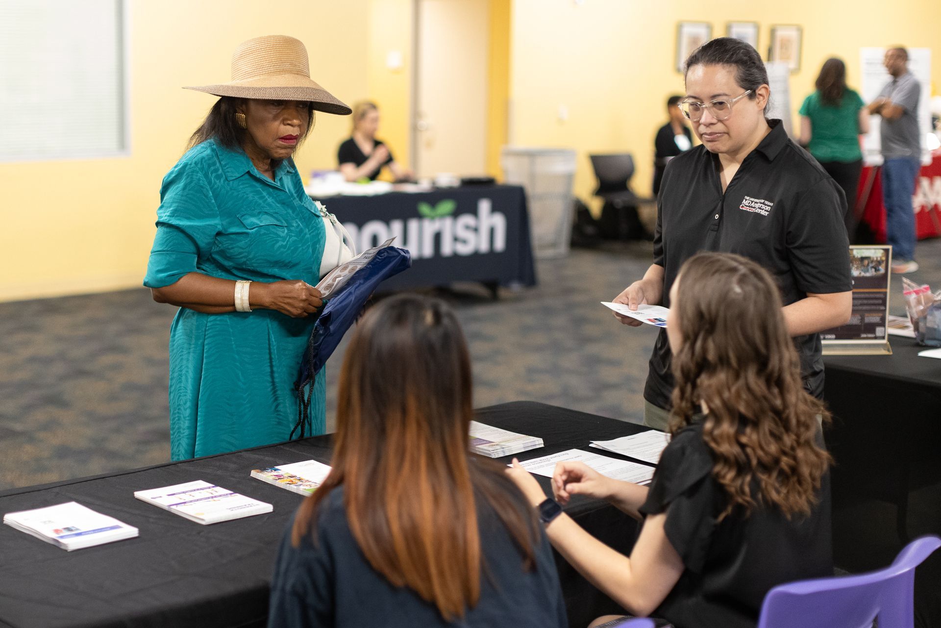 People at tables with informational materials at an event. Woman in turquoise dress and straw hat speaking to people.