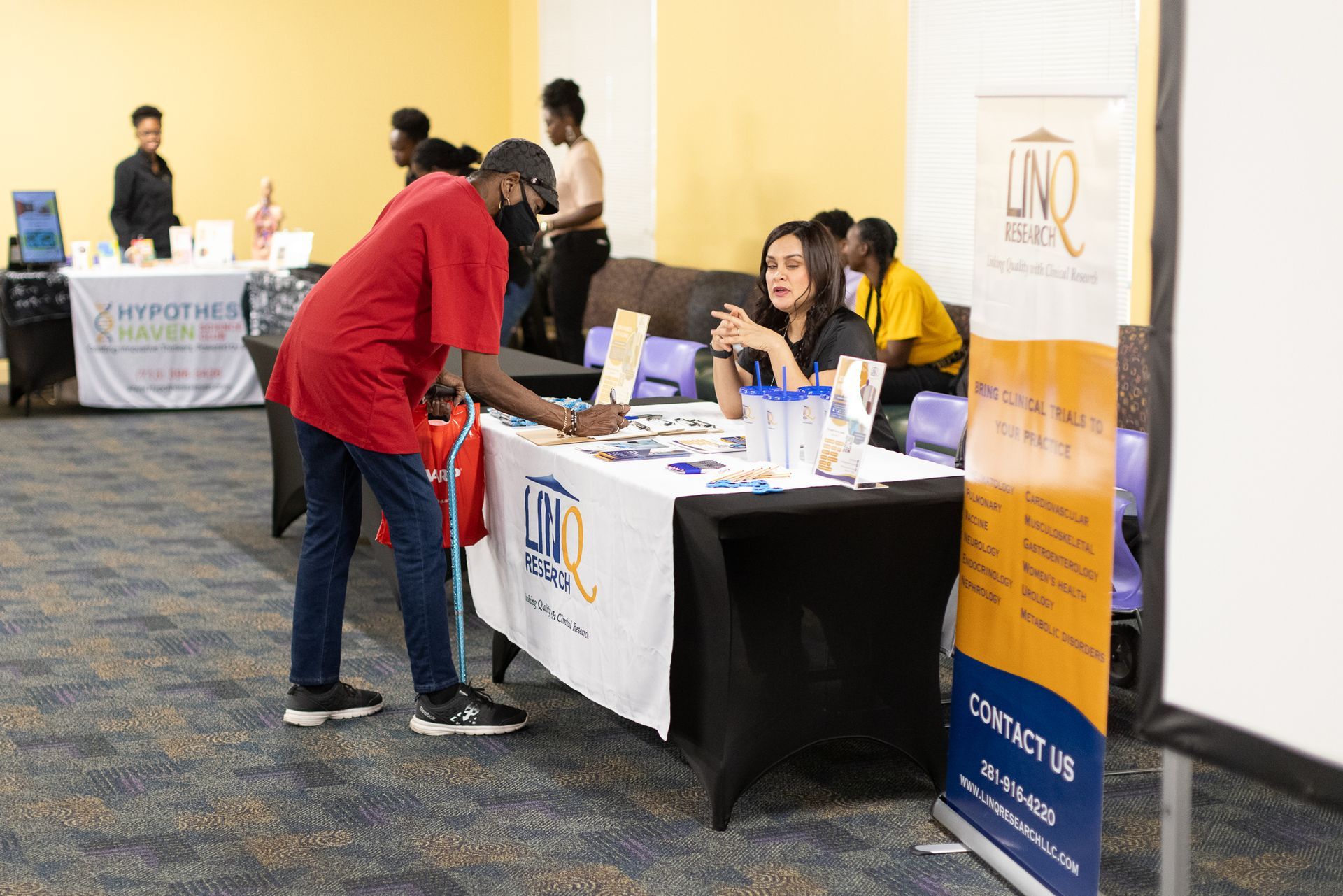 People at tables with promotional materials at a fair. One person in a red shirt at a table.