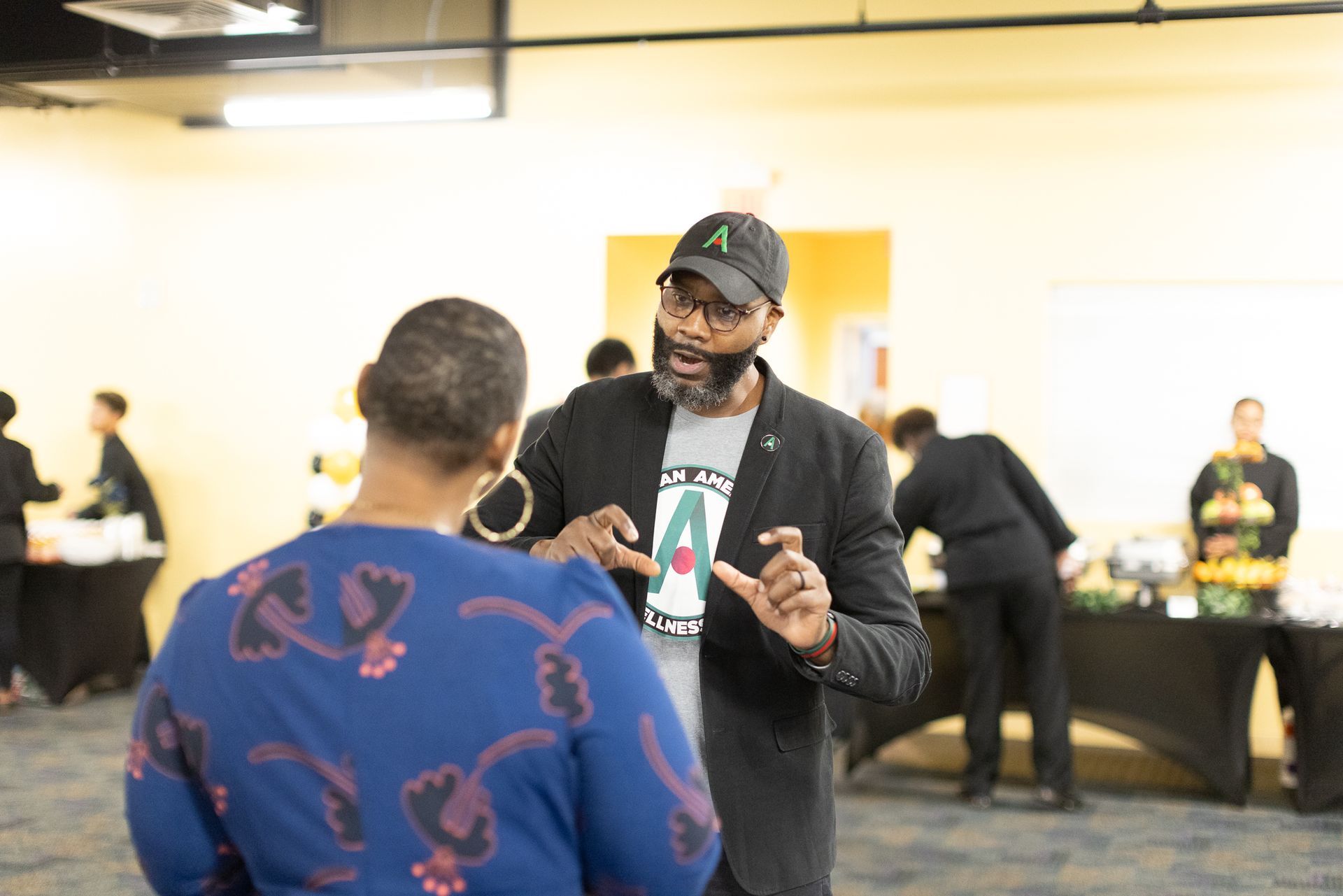 Two people conversing in an event space. Man in a cap and tee gestures with hands, speaking to a woman.