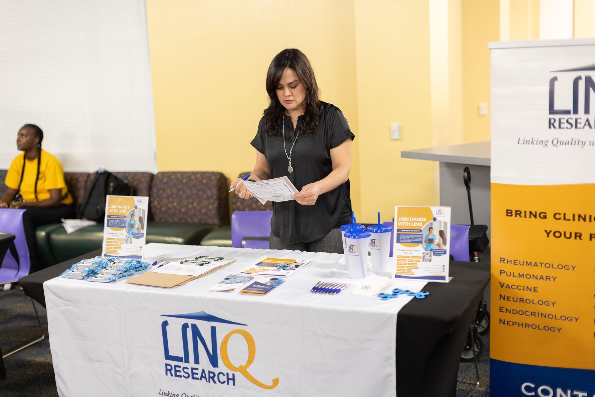 Woman at table for LinQ Research, arranging brochures. A banner and giveaways are also visible.
