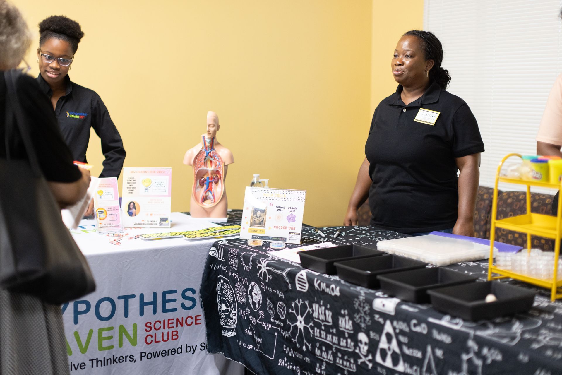 Two women at a science club booth, one assisting a customer. Table displays anatomy model and science kits.