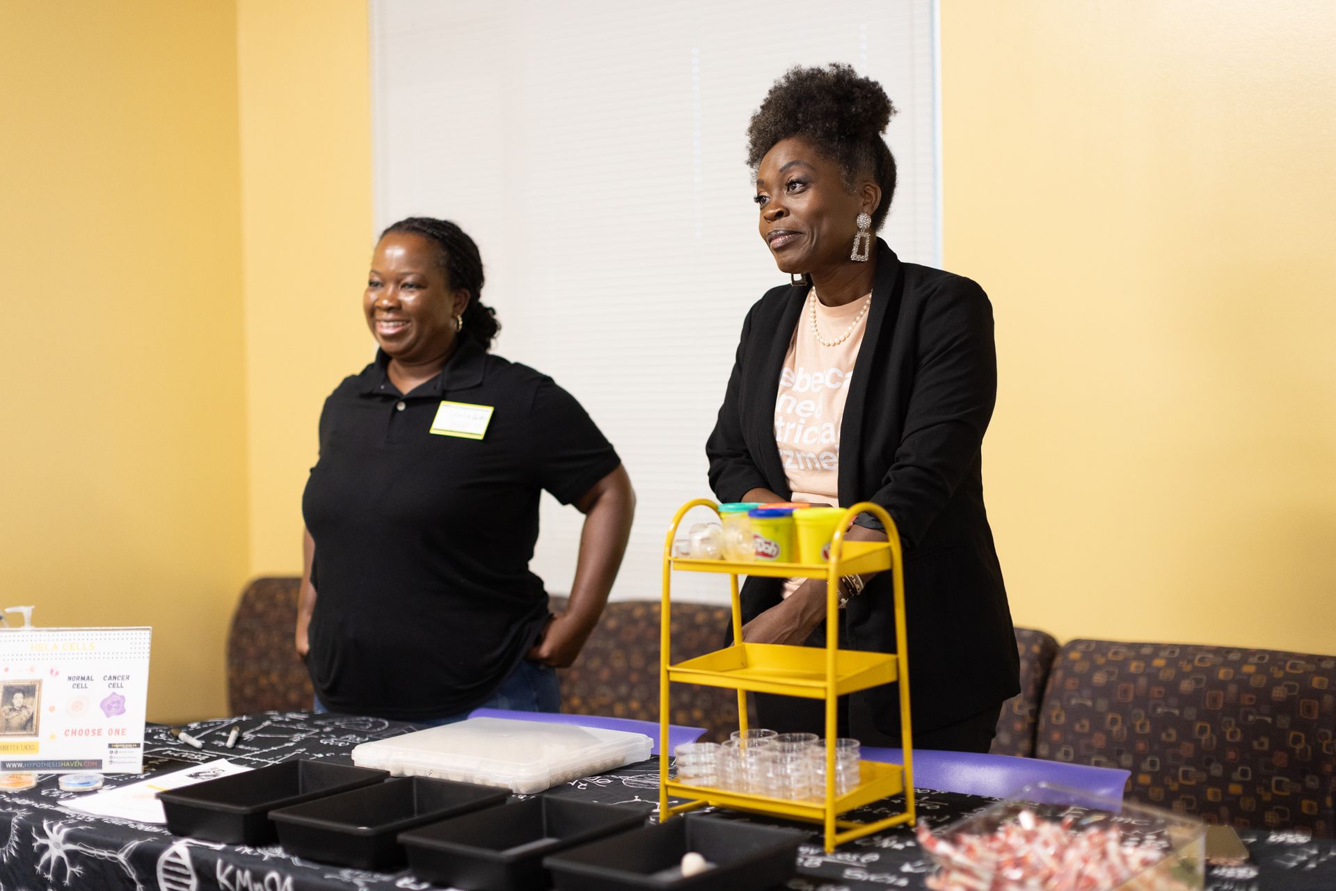 Two women standing behind a table with food, one wearing a black blazer, the other a black t-shirt.