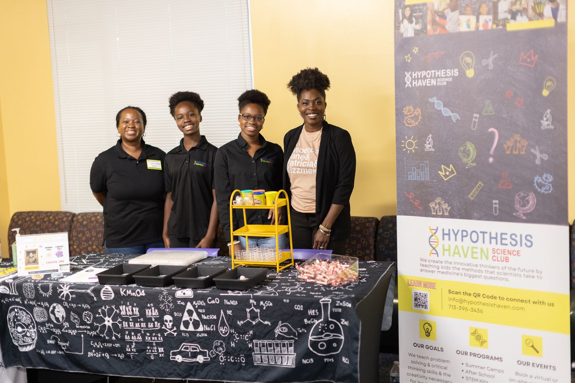 Four people stand behind a table with science supplies and a banner for 