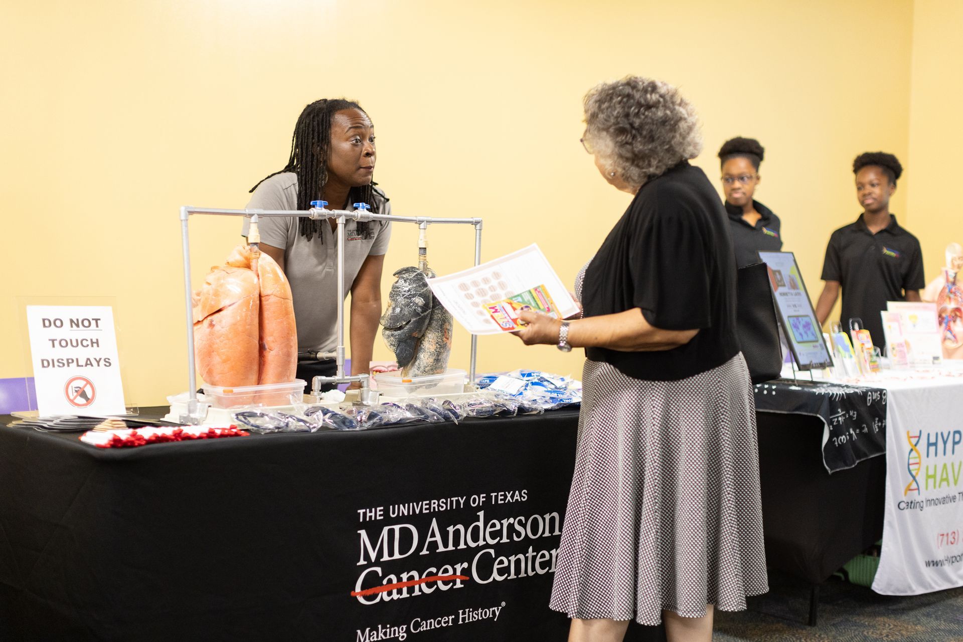 Person speaking to a woman at a cancer center information booth, with models of lungs and educational materials on the table.