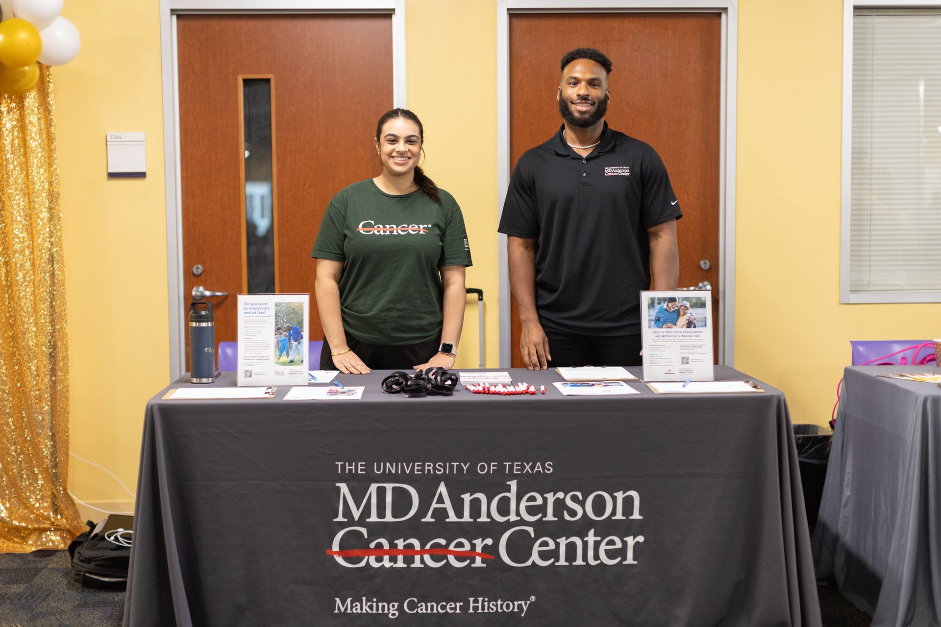 Two people stand behind a table with an MD Anderson Cancer Center logo, promoting cancer awareness.