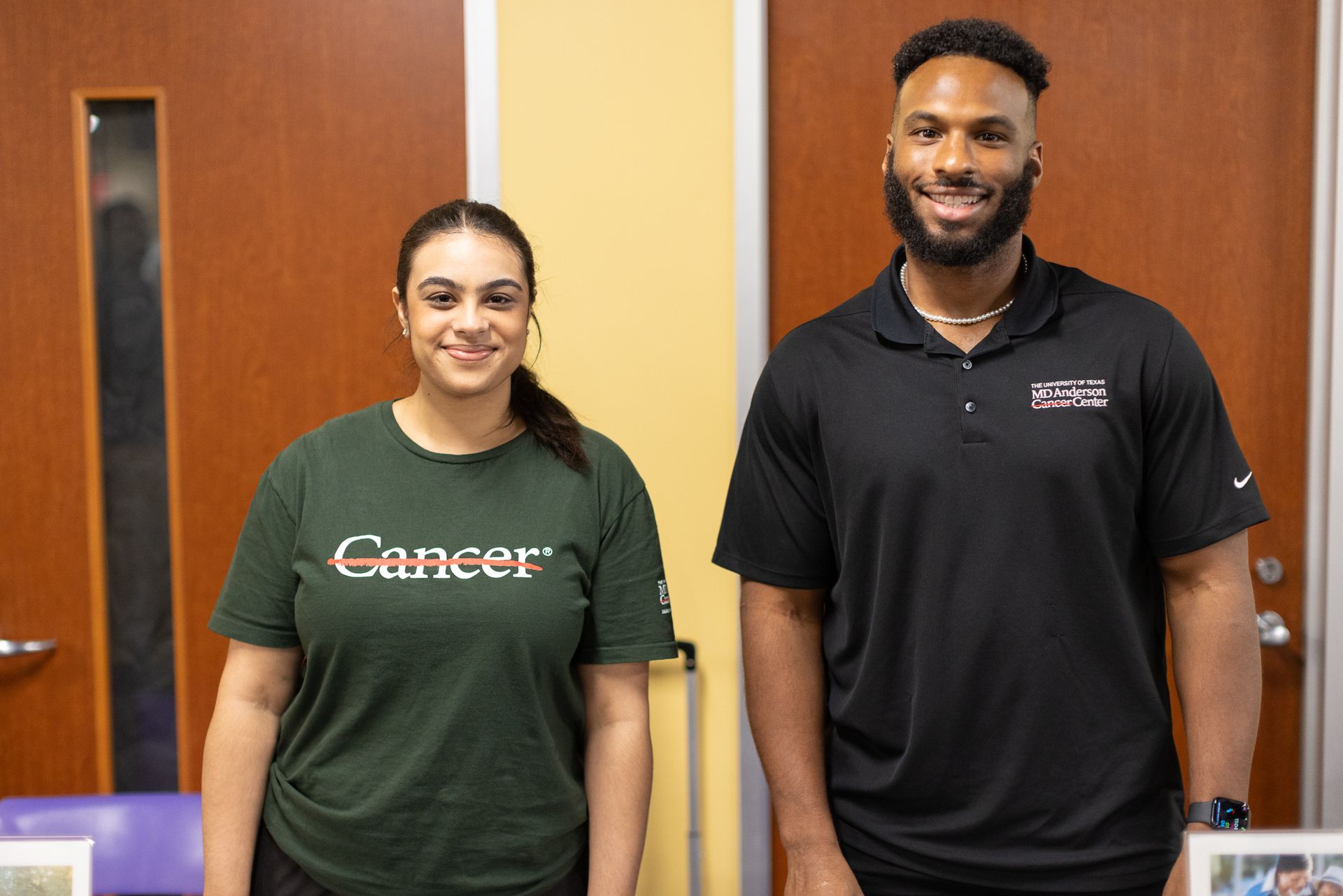 Woman in green shirt, man in black shirt, stand in front of wooden doors. Both smile.
