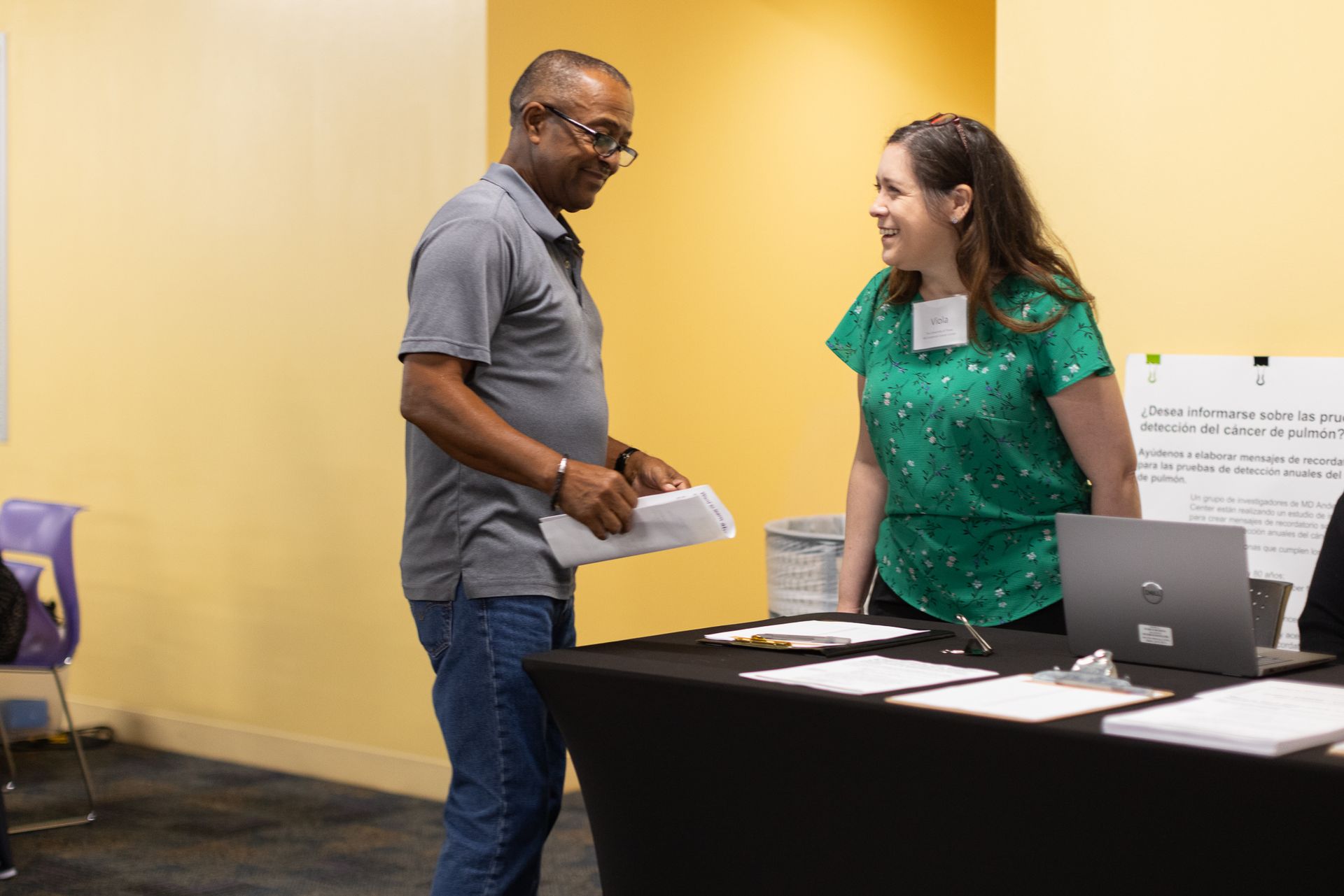 Man and woman talking at a table with papers and a laptop in a room with a yellow wall.