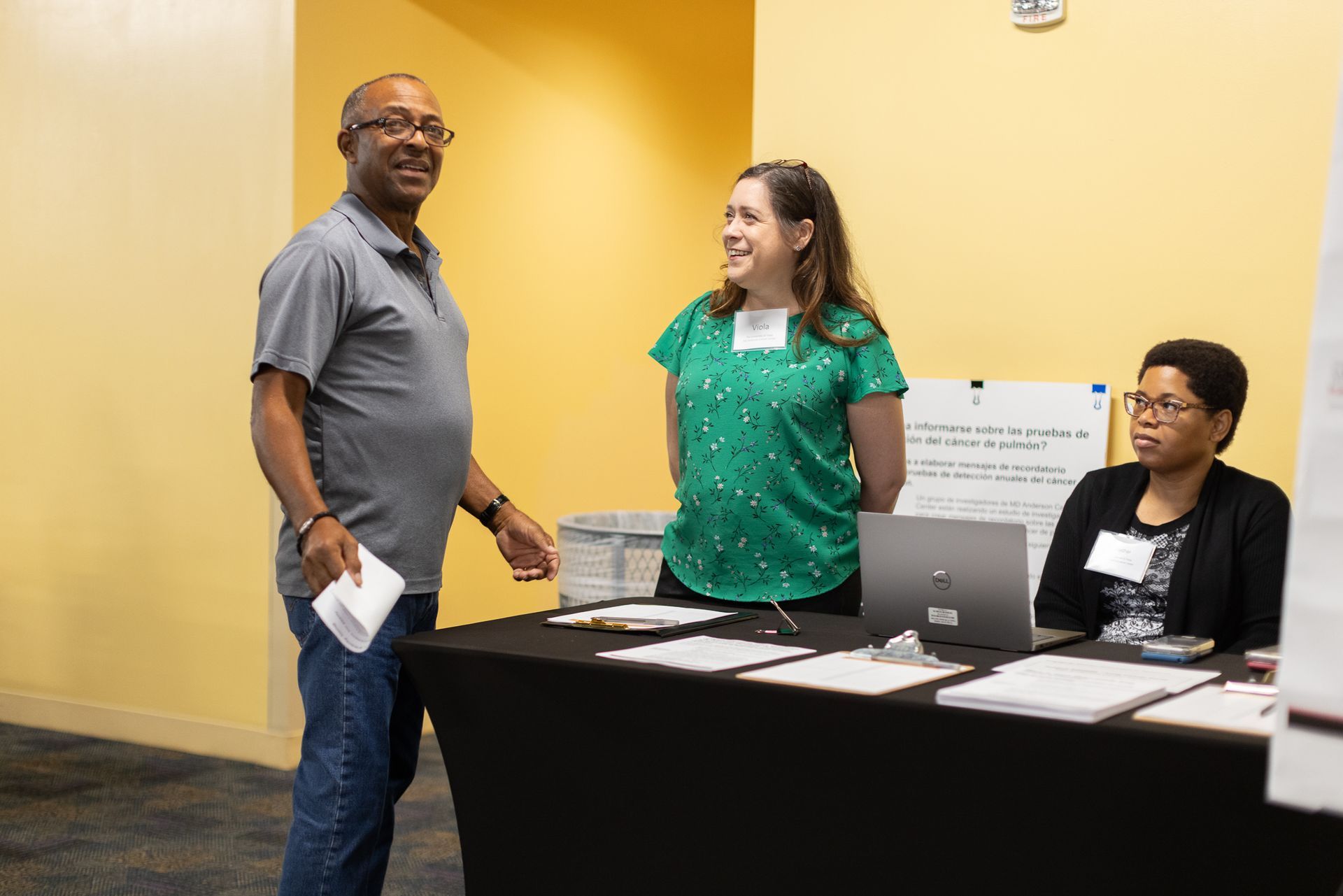 Man speaking with two people at a registration table. Yellow wall, black tablecloth, laptop.