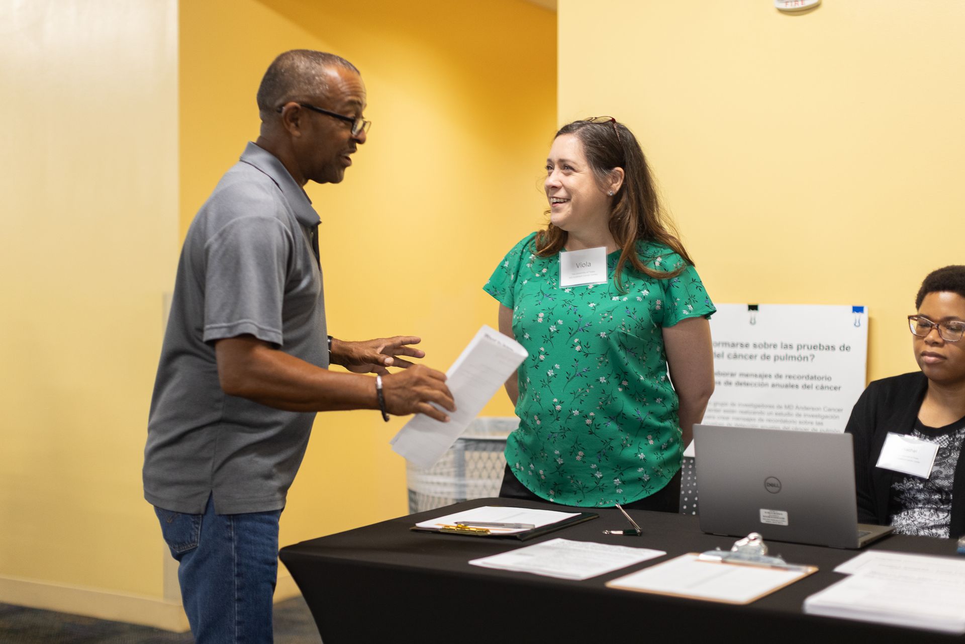 Man at a table hands papers to a woman in a green shirt. A third person sits at the table. Yellow wall.