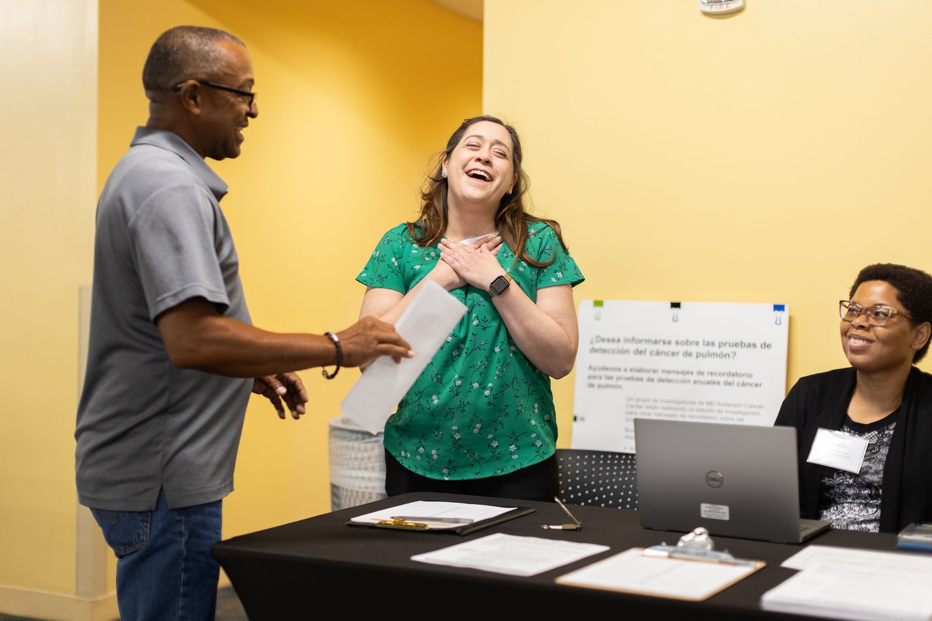 Man hands papers to woman laughing at table, other woman smiles nearby. Yellow wall, table with laptop.