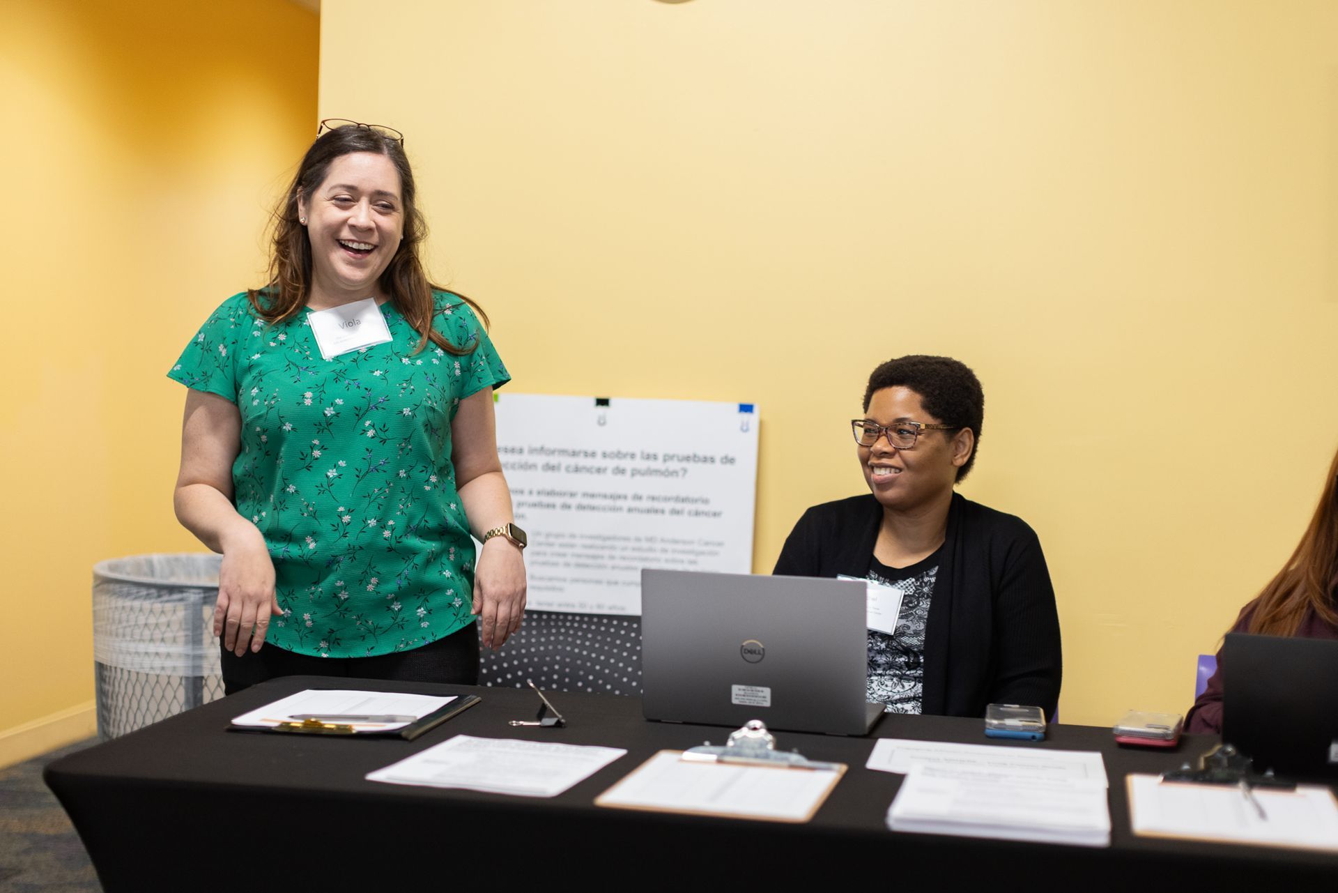 Two women at a table with papers, one in green shirt smiling, the other working on a laptop. Yellow wall in background.