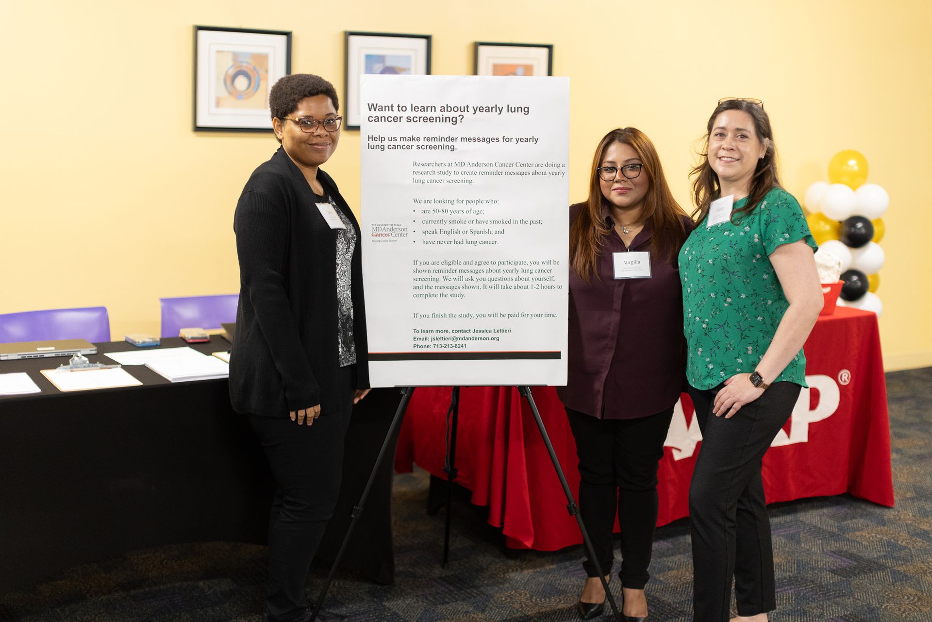 Three people stand near a poster on an easel. A table with brochures is in the background. The scene is brightly lit.