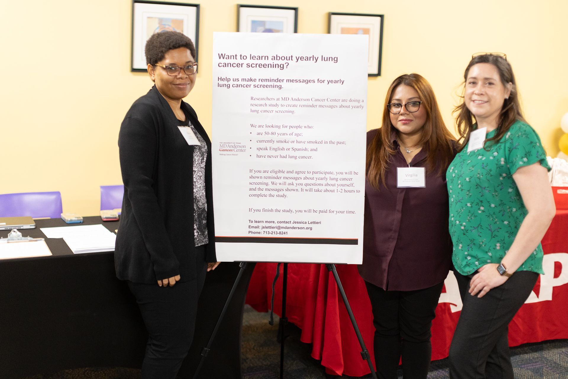 Three people stand next to a poster. They are smiling at a table with a red cloth. The background is a room.