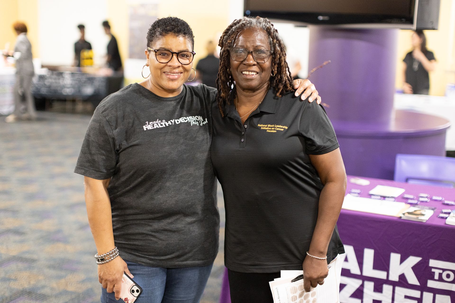 Two smiling people with arms around each other at an event, purple signage in the background.