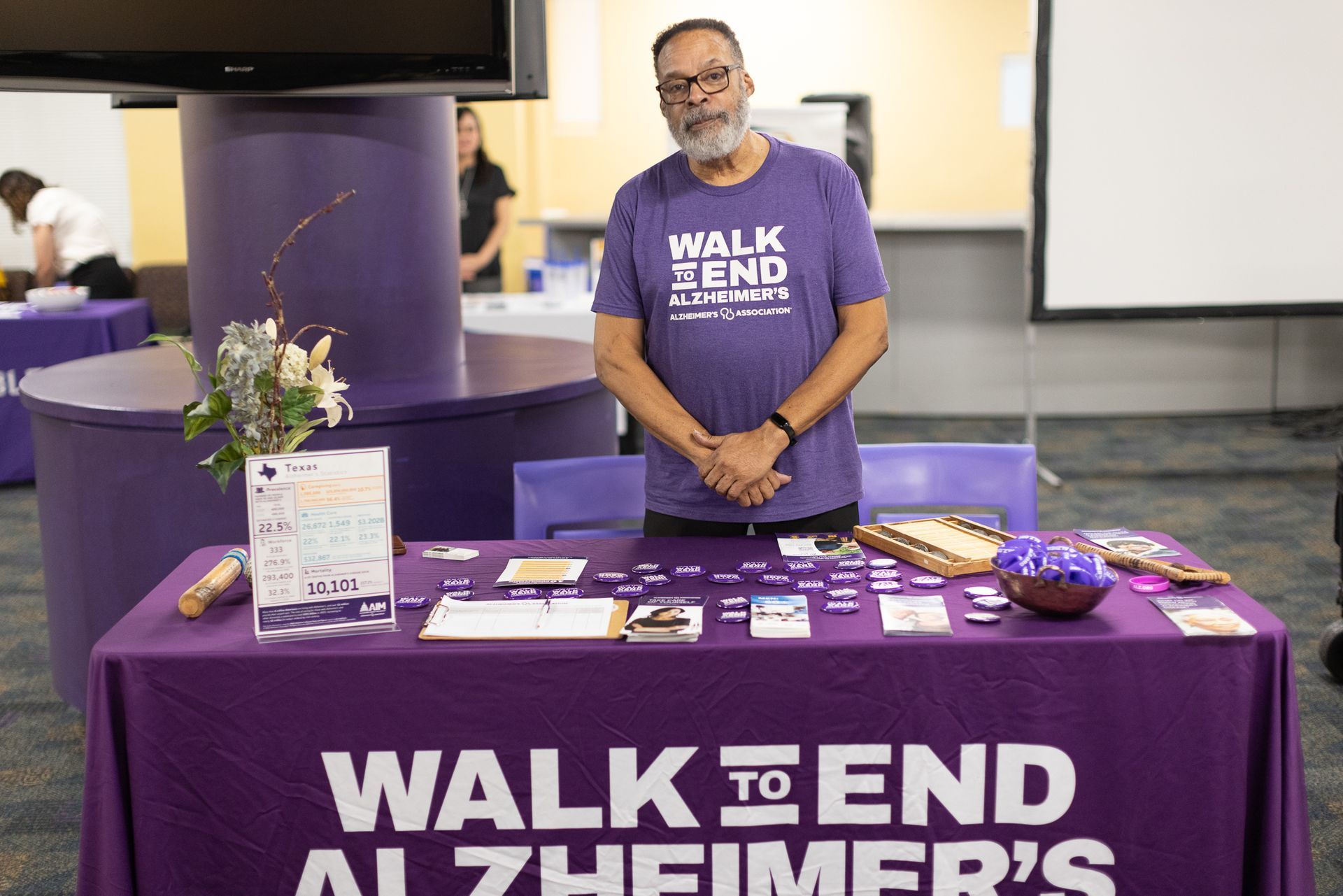 Man at a table promoting Walk to End Alzheimer's, wearing a purple t-shirt. Table with informational materials.