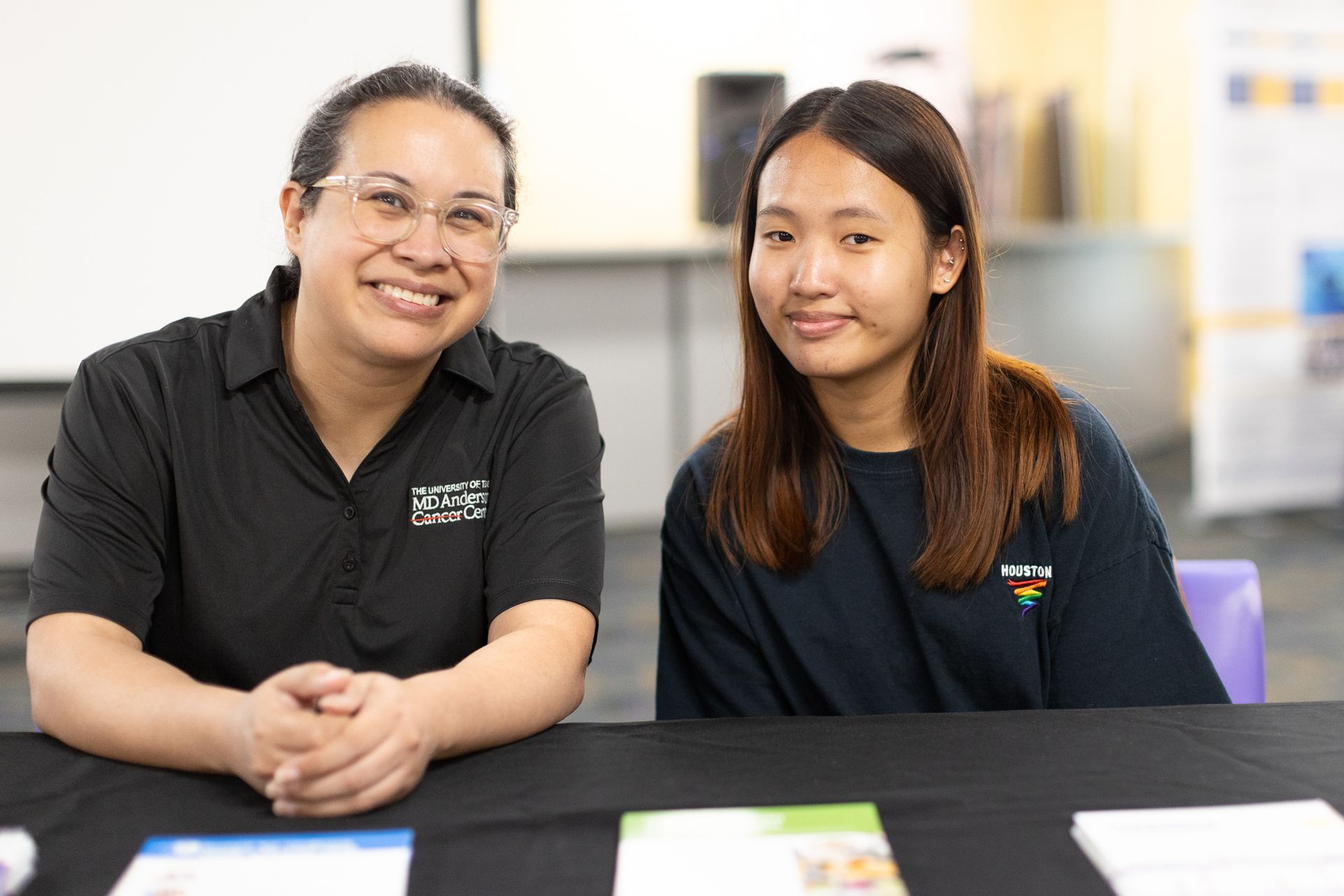 Two people seated behind a table, smiling. One wears a black shirt, the other a navy blue shirt.