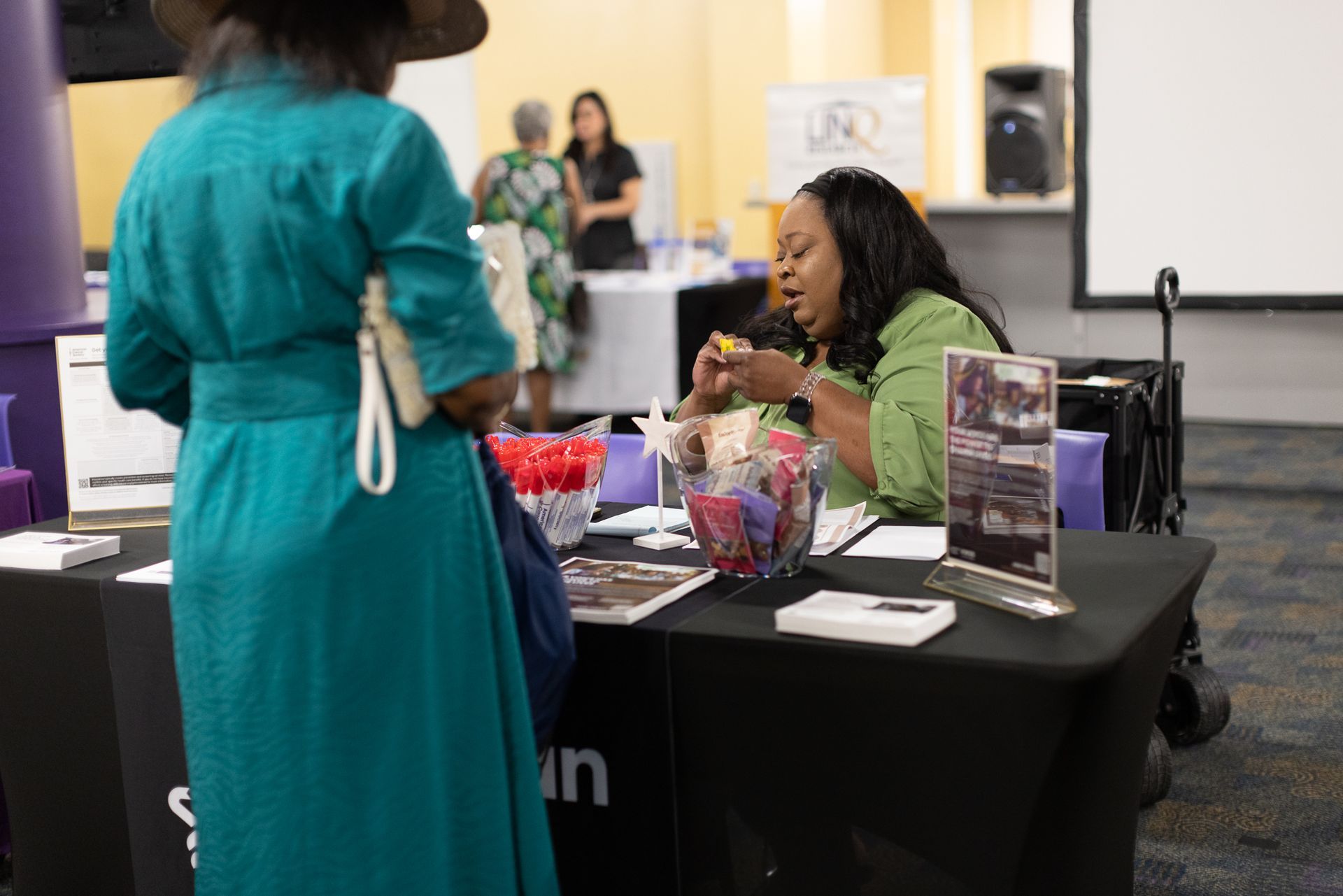 Person at a table with promotional materials talks to a visitor at a conference.