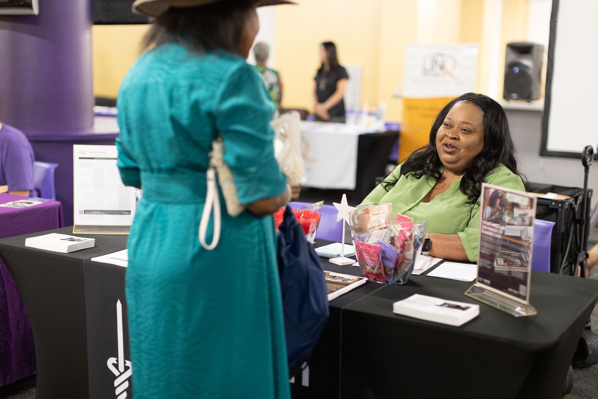 Woman in teal dress at a table talks to another woman at an event.