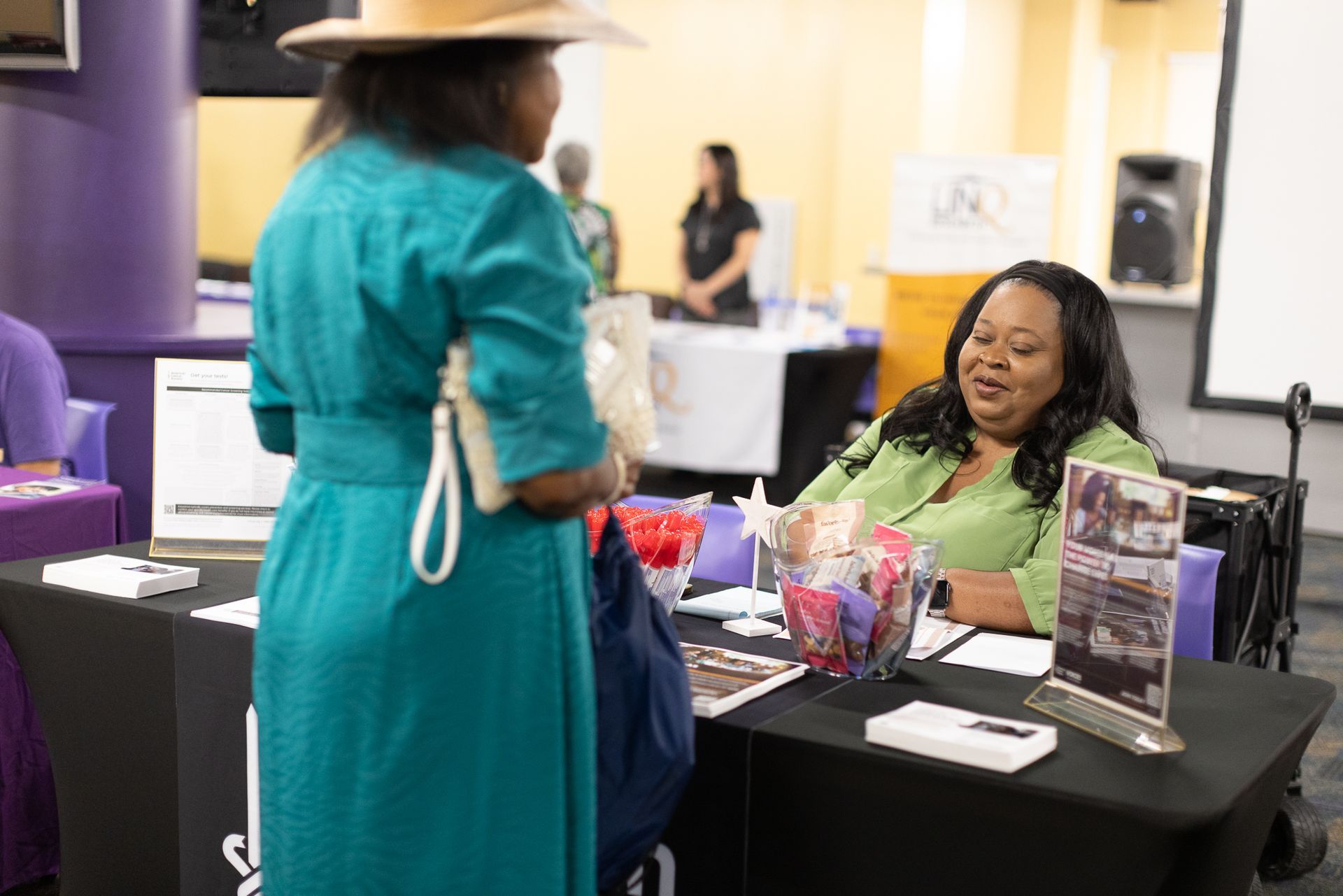 Two women at an event table, one speaking and the other sitting. Table has brochures and a sign.