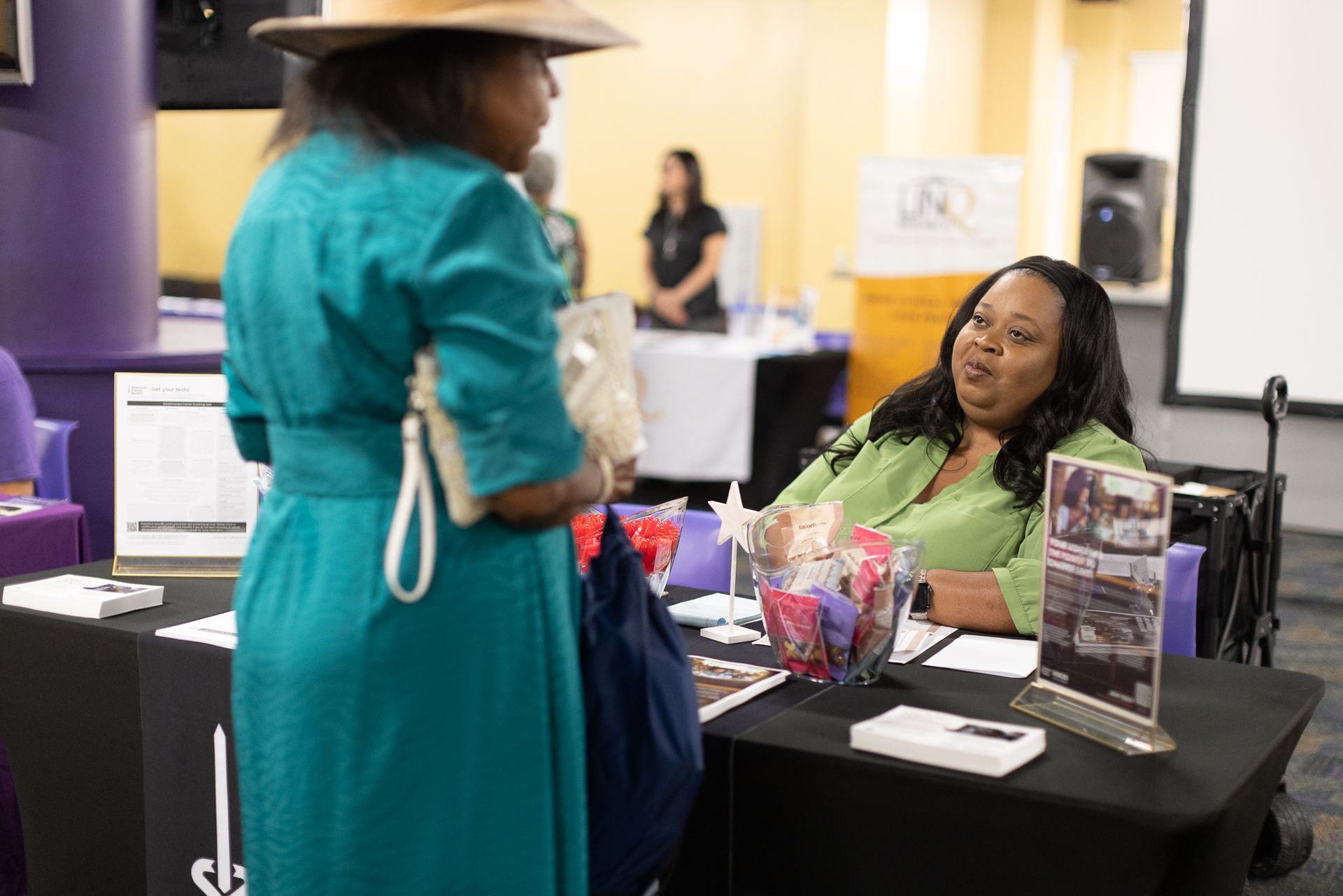 Woman in green dress talks with another woman at a table with promotional items.