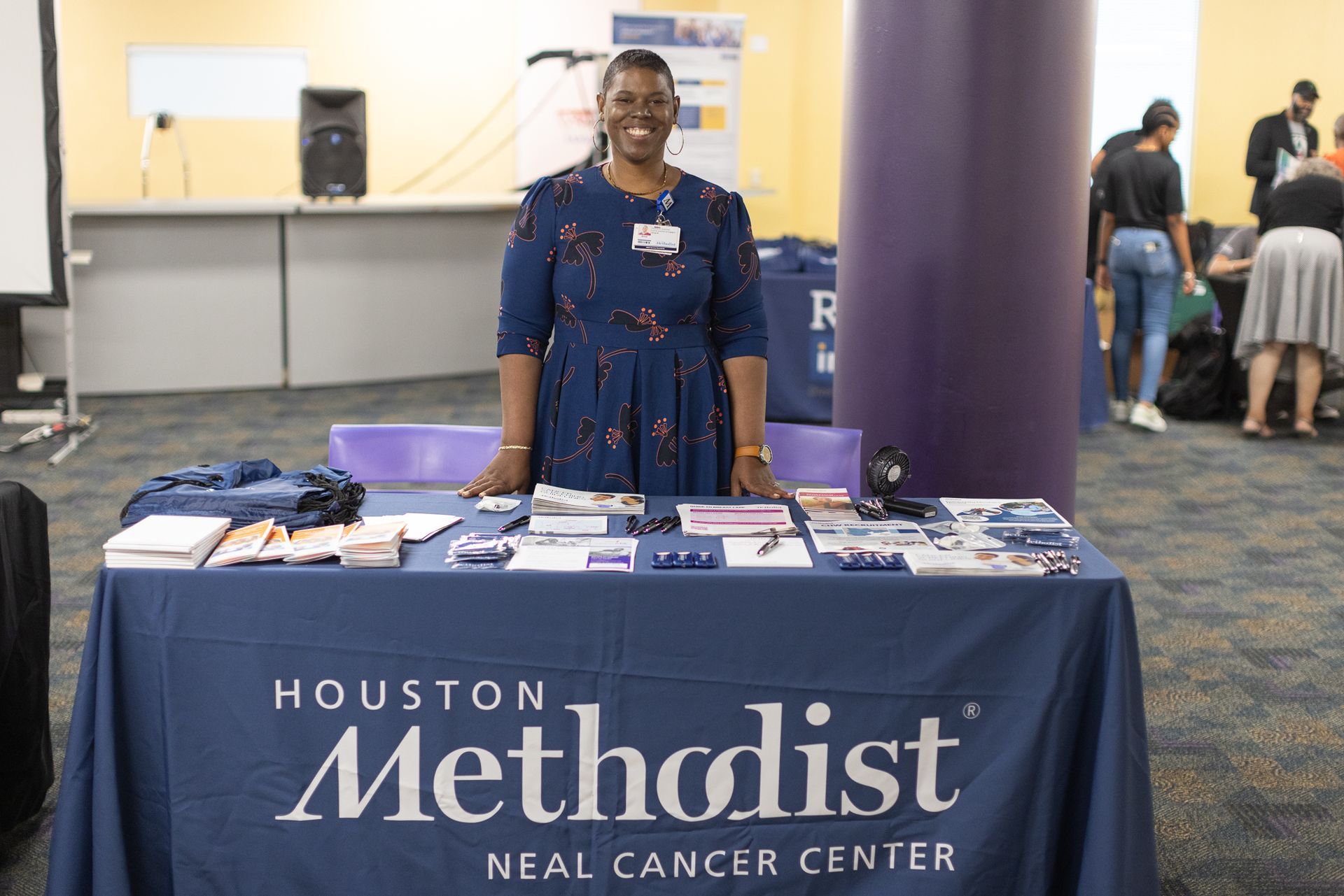 Woman at Houston Methodist Neal Cancer Center table, smiling. Blue table with brochures, flyers, and logo.