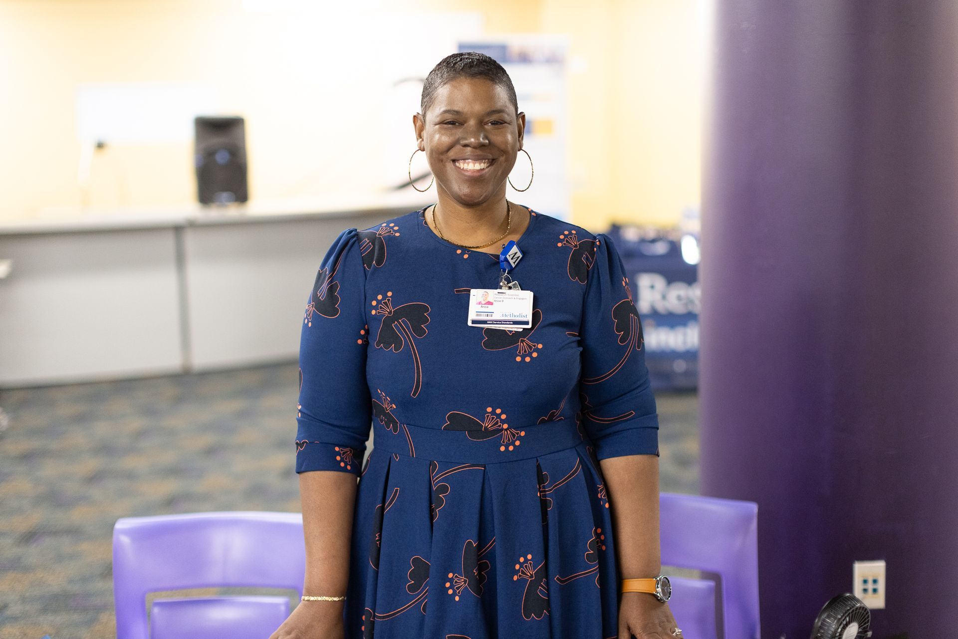 Woman in a navy patterned dress smiles, stands behind purple chairs, and near a purple pillar.