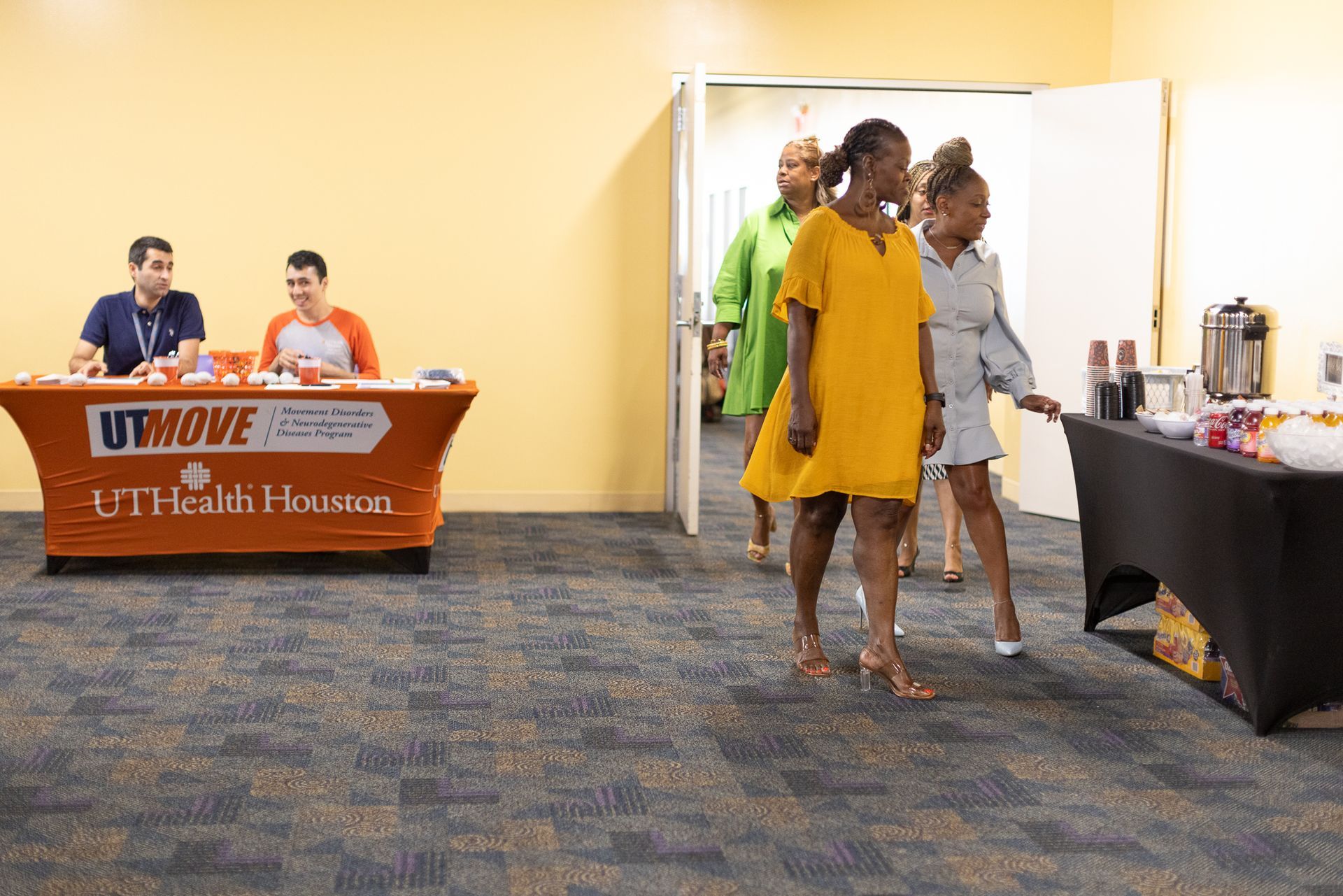People walk past a table with refreshments and a table with two seated people at a presentation.