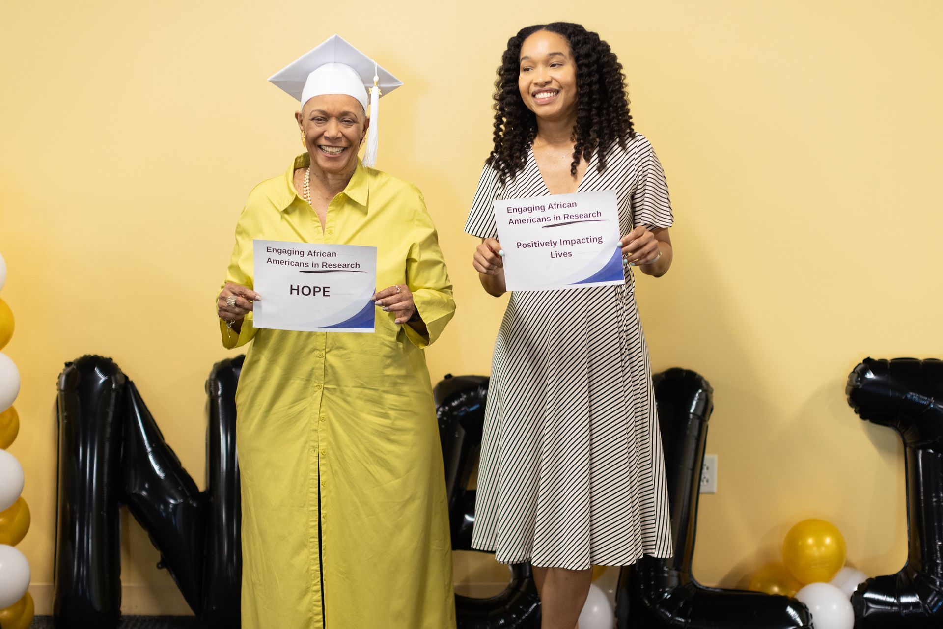 Two women holding certificates, posing in front of a yellow wall with balloons and black letters that spell out NLJ.