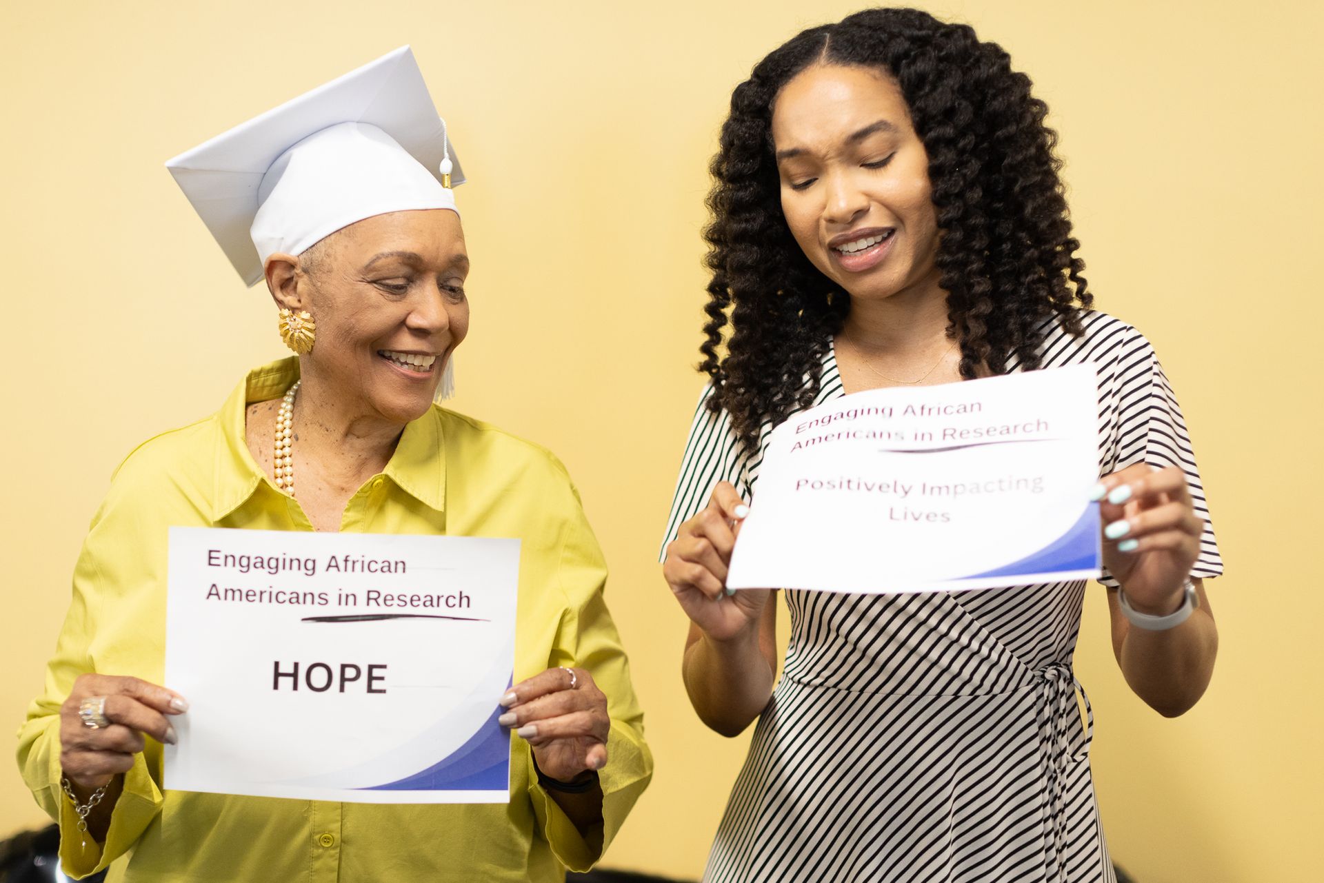 Two women holding signs. Older woman in a graduation cap smiles. Younger woman points at a sign.