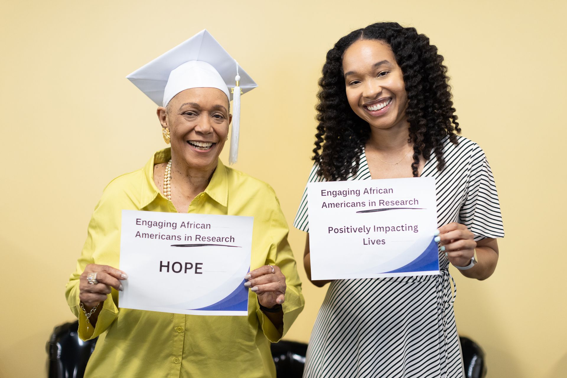 Two smiling women holding signs, one in a graduation cap, both smiling; yellow background.