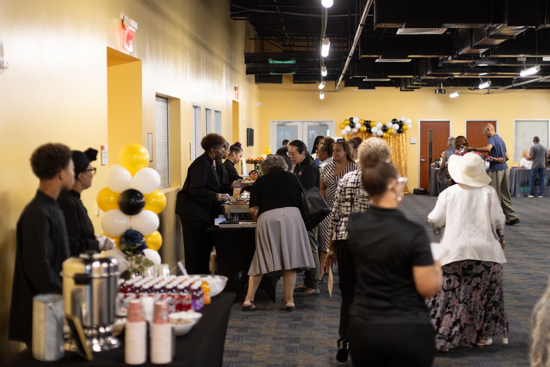 People at an event with food and drinks. Yellow and black balloons.
