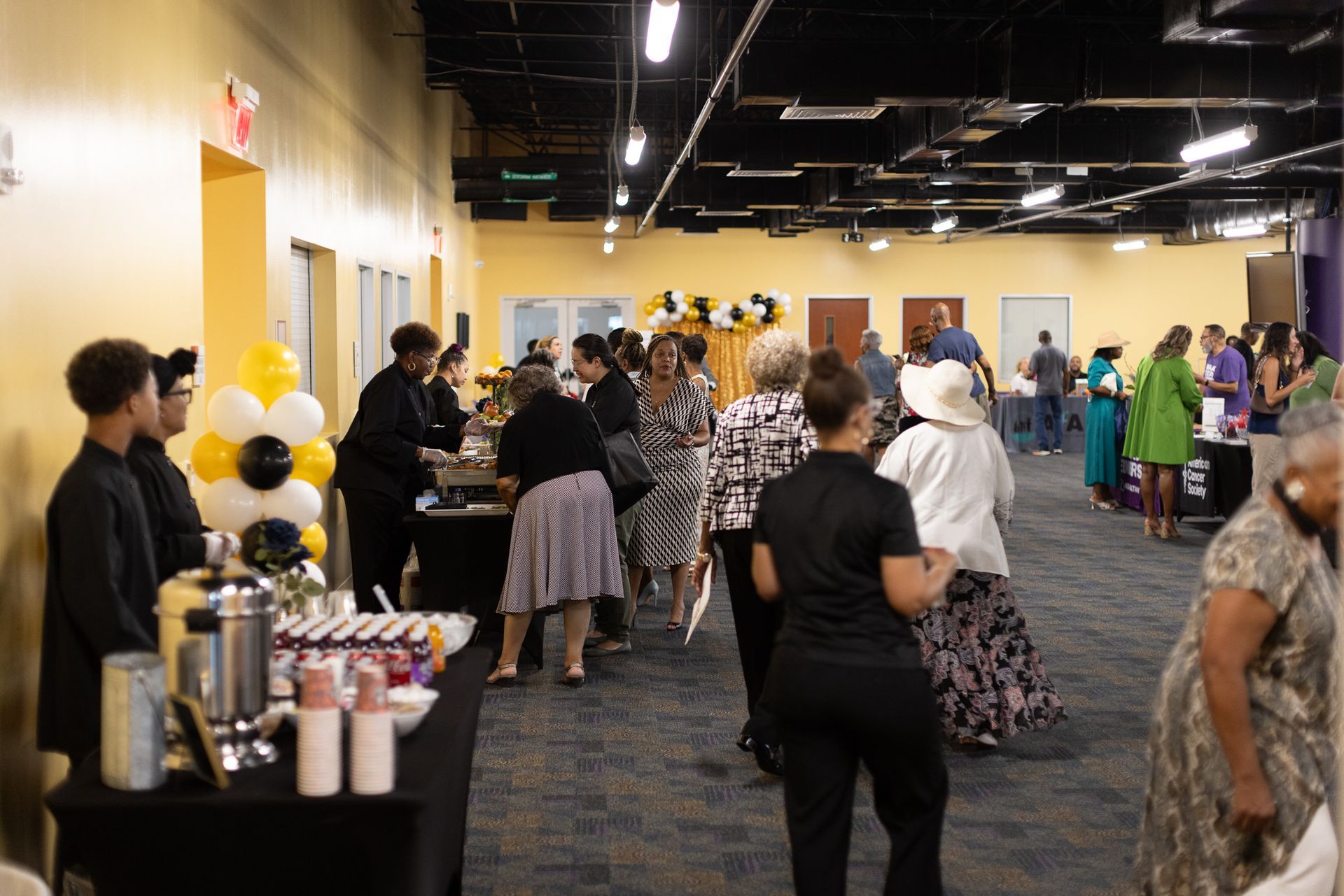 People at an event with food and drinks. Yellow and black decorations, purple and yellow walls.