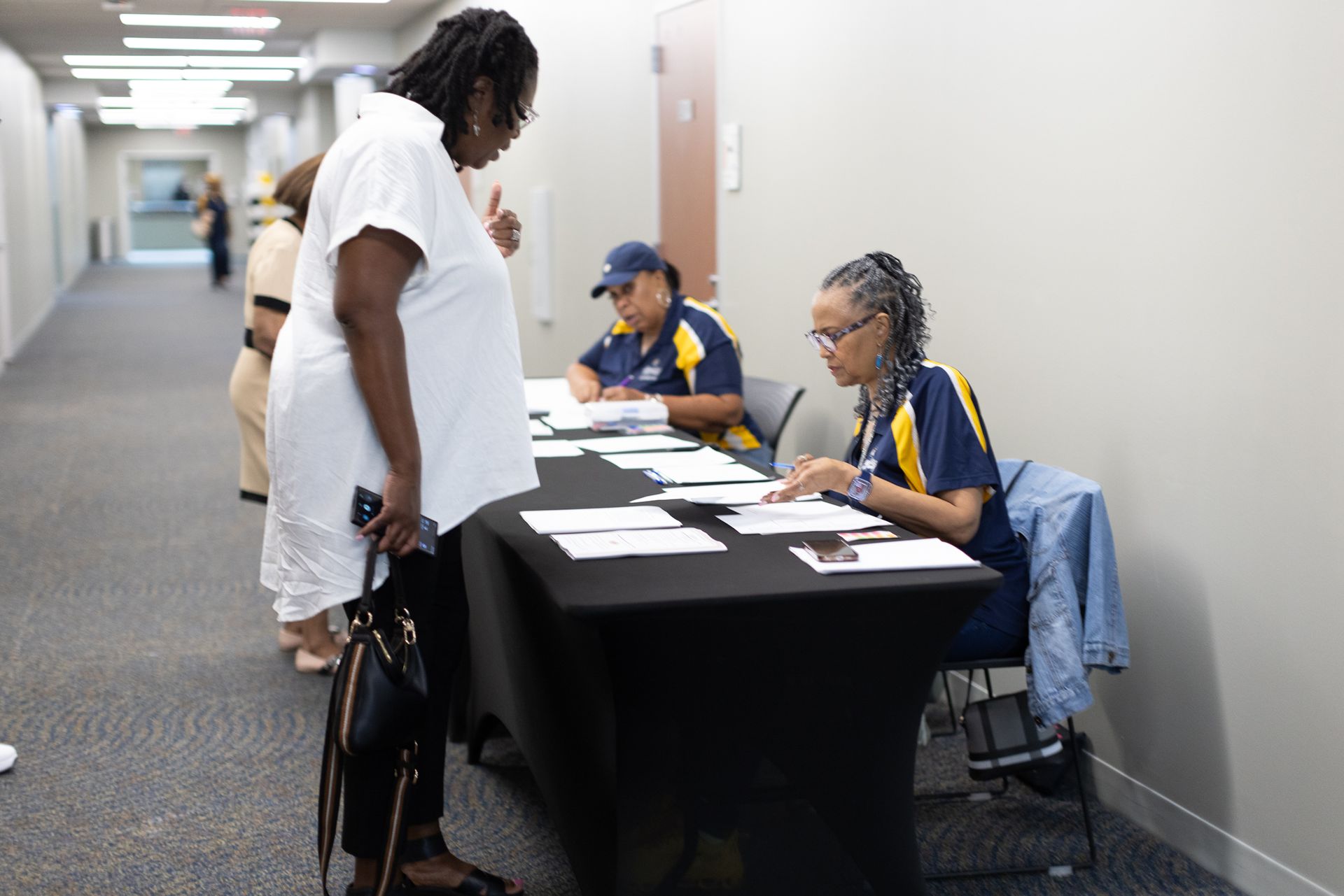Woman at a table checking in, two people sitting behind the table, and person in hallway.