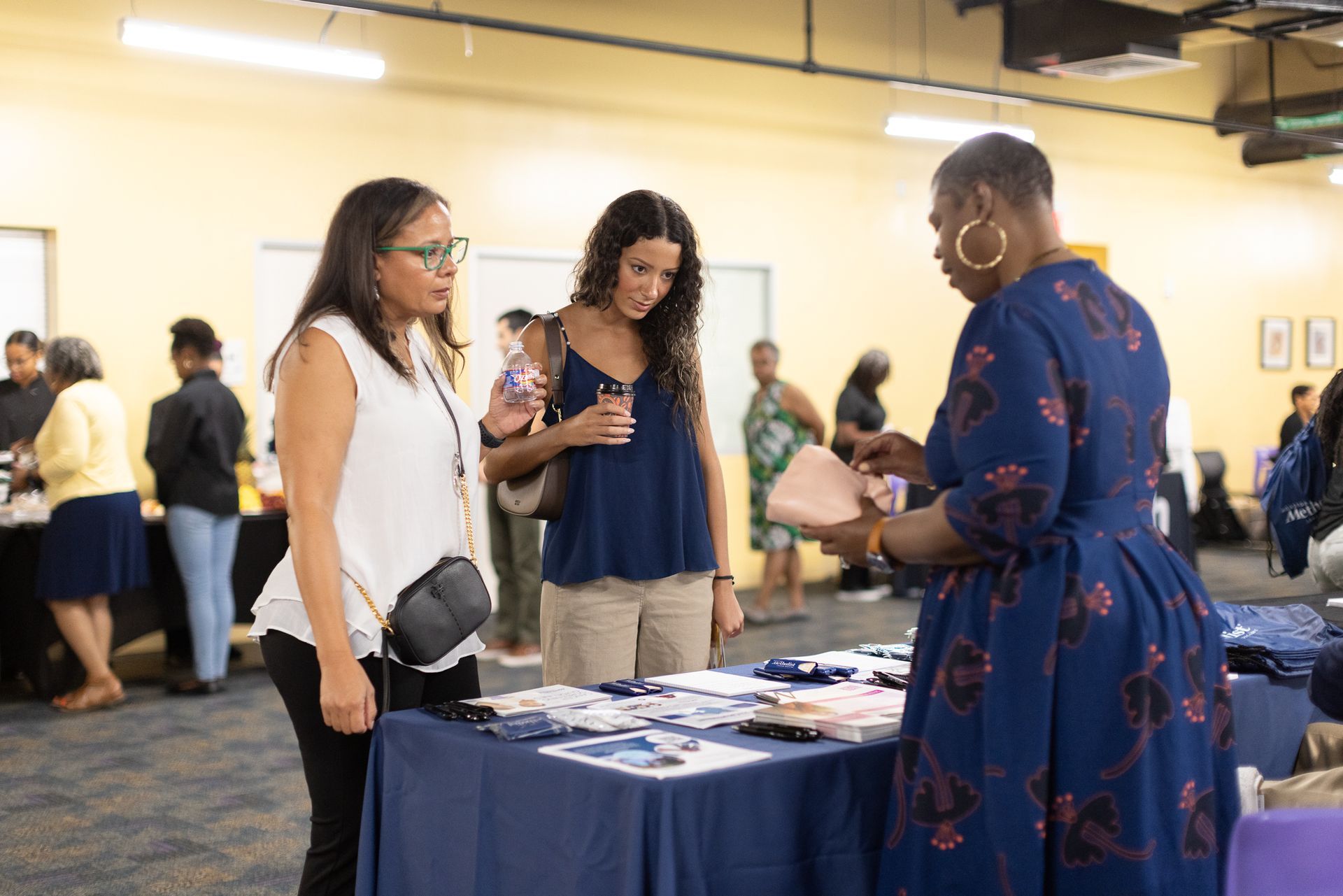 Three women at a table with promotional items; a woman in a blue dress is showing items to two others.