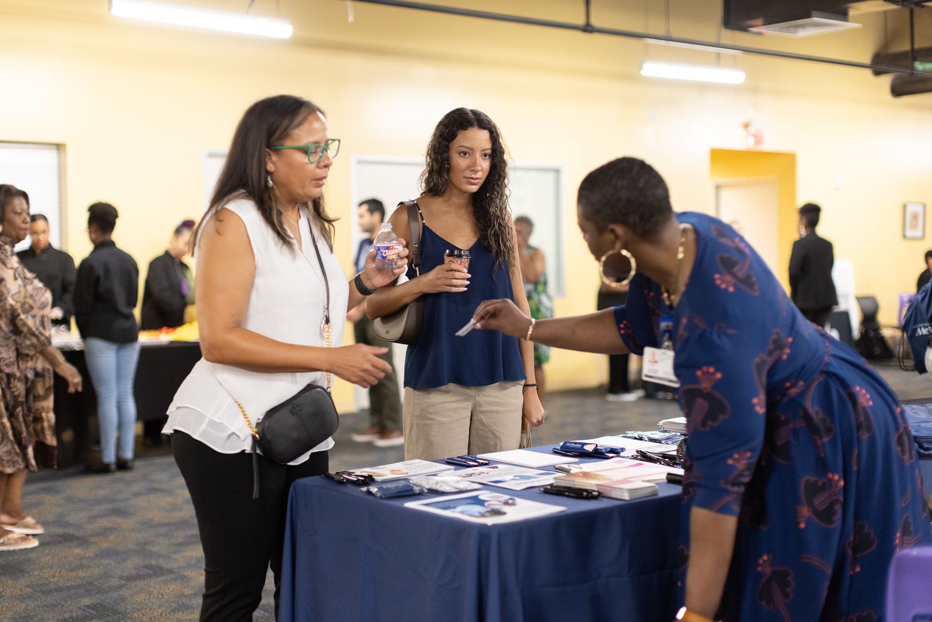 People at a table, discussing materials. One woman hands a card to another. Other people stand around in a room.