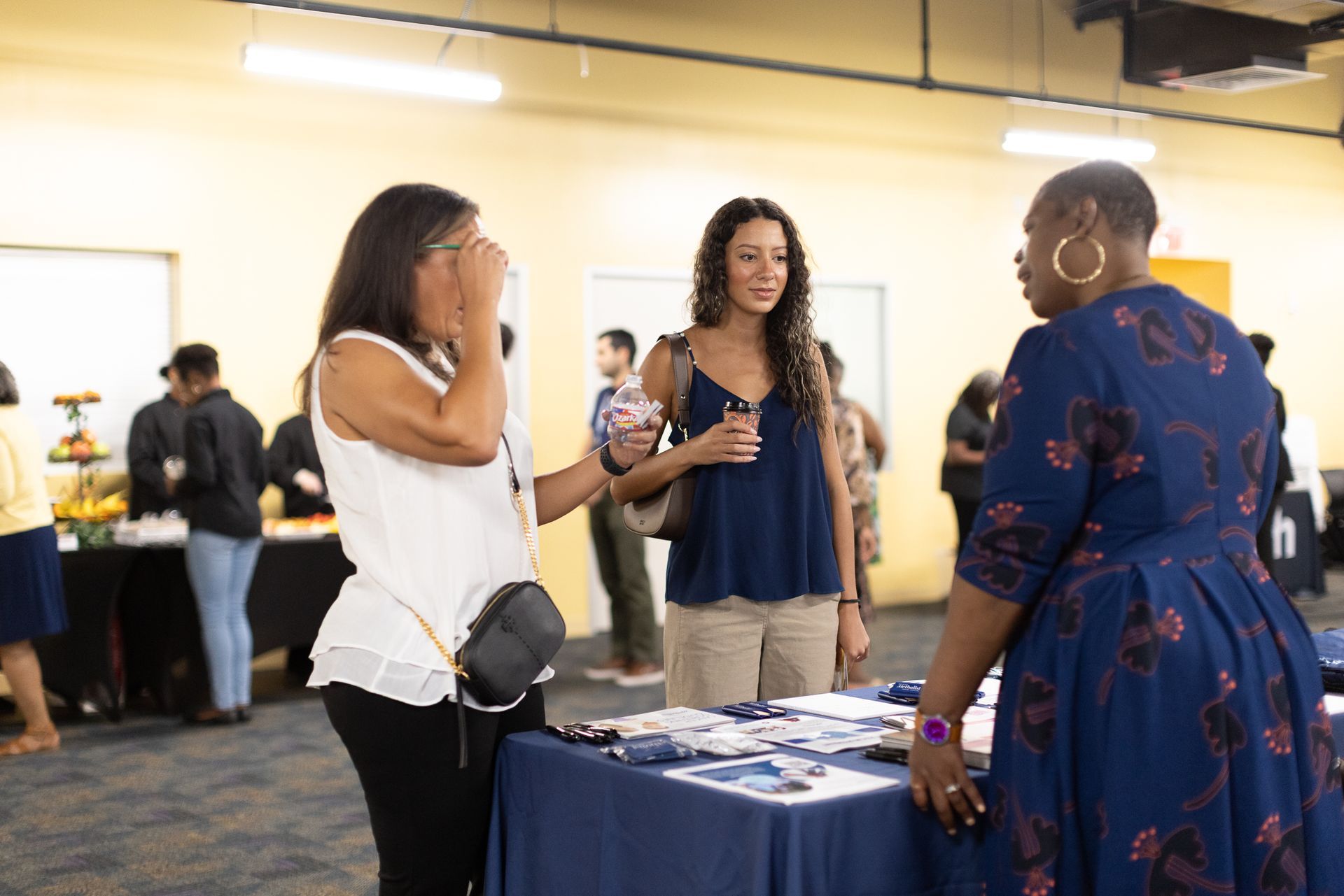 Three people talking near a table with materials in a room with other people.