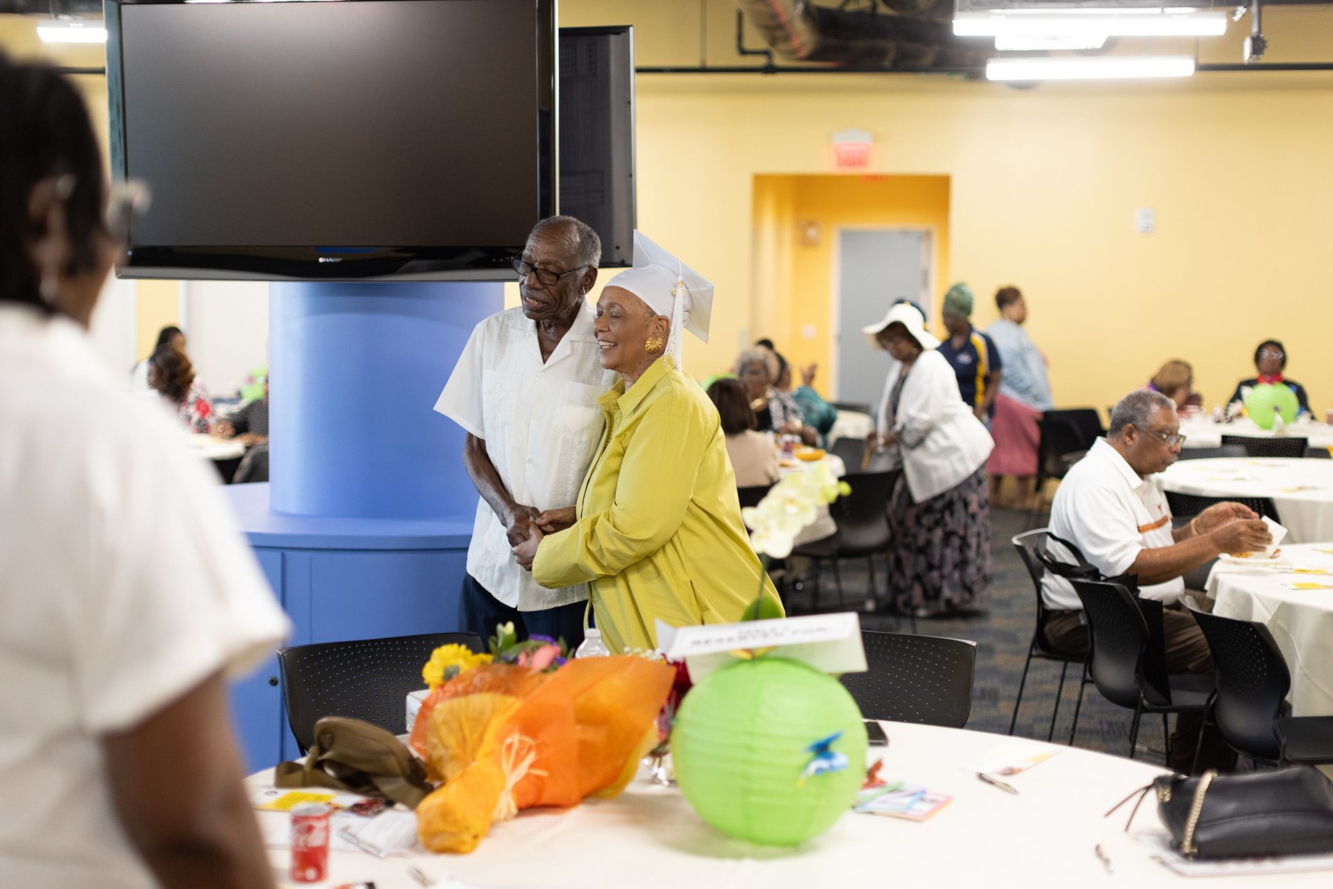 Couple smiling, arm in arm, in a community center; others at tables nearby.
