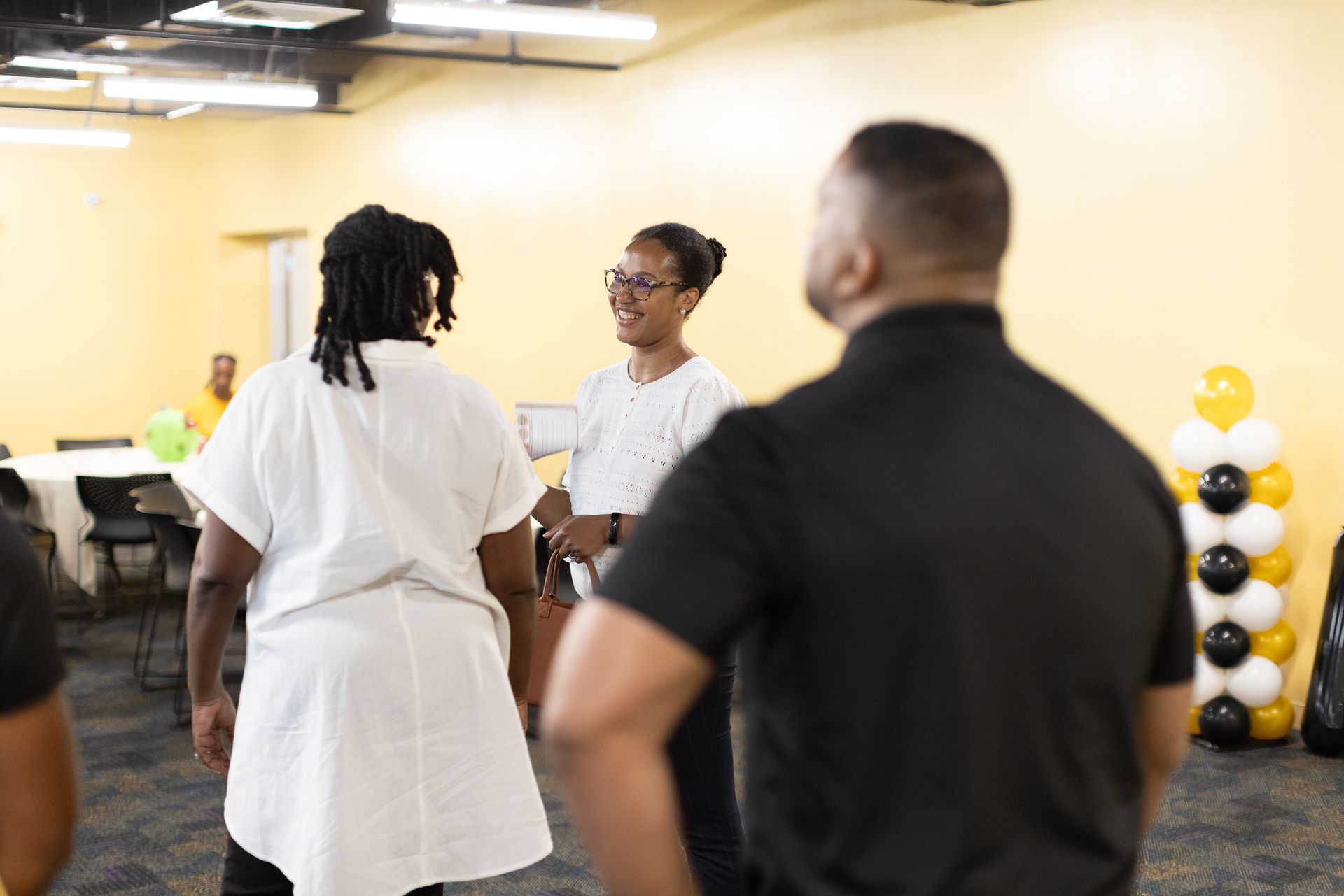 People in a room talking. Woman in white shirt smiles at two others. Balloons in corner.